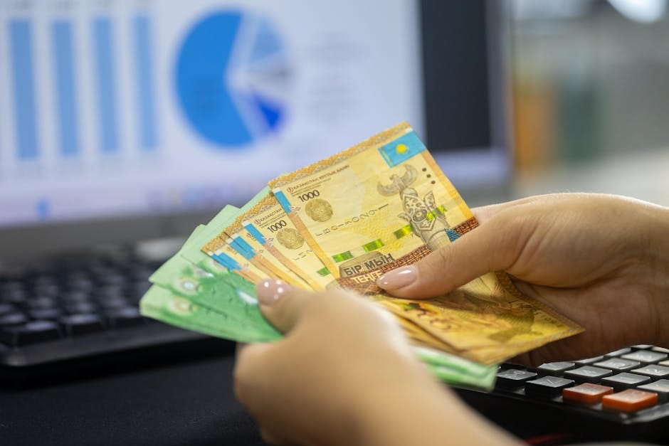 Manos humanas contando billetes de tenge kazajo cerca de una computadora portátil que muestra gráficos financieros.