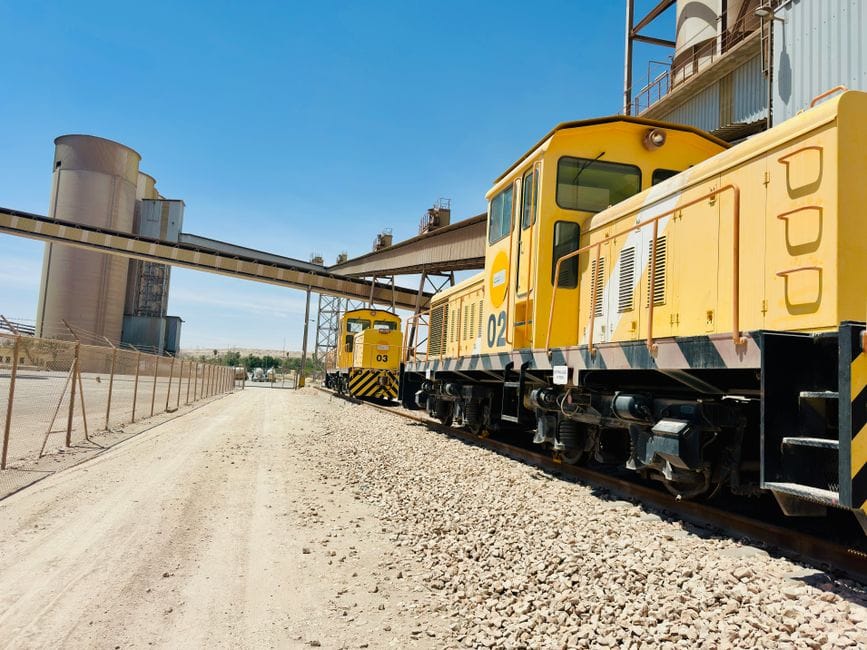 Trenes industriales amarillos en una vía férrea junto a una fábrica de cemento en un día despejado.