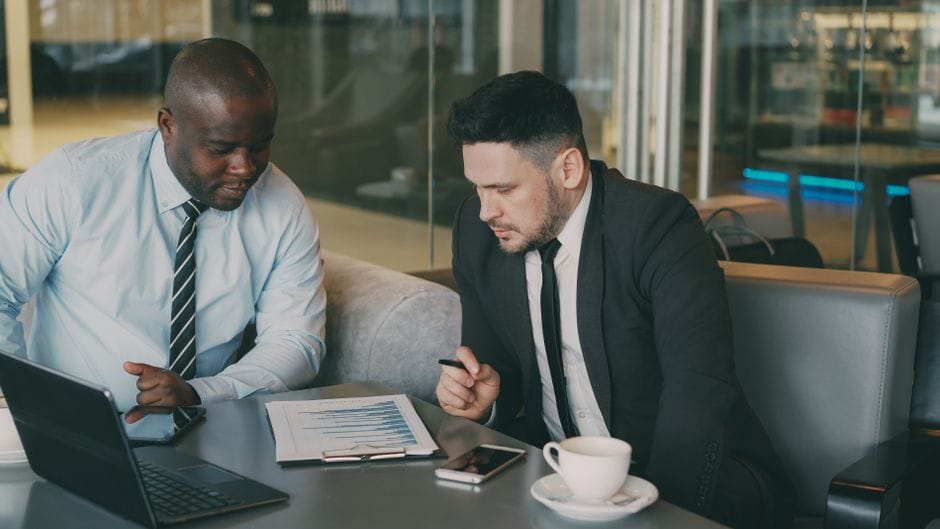 Dos empresarios revisando un informe financiero con su computadora portátil y café en un entorno de oficina.