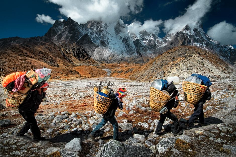 Porteadores transportando mercancías a través del escarpado terreno del Himalaya en Nepal, con un impresionante paisaje montañoso de fondo.