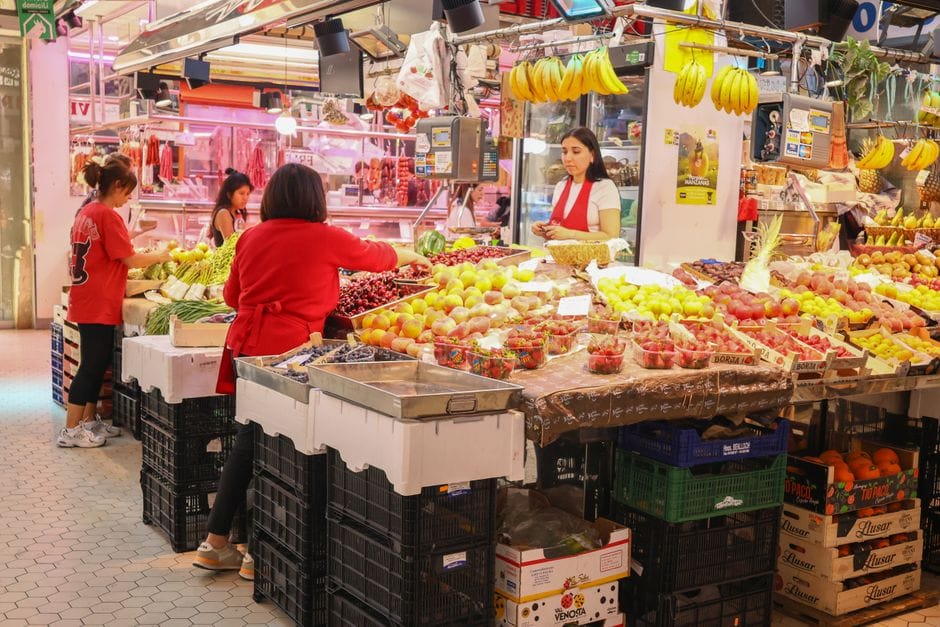 Escena colorida de un mercado cubierto con compradores adquiriendo frutas y verduras frescas.