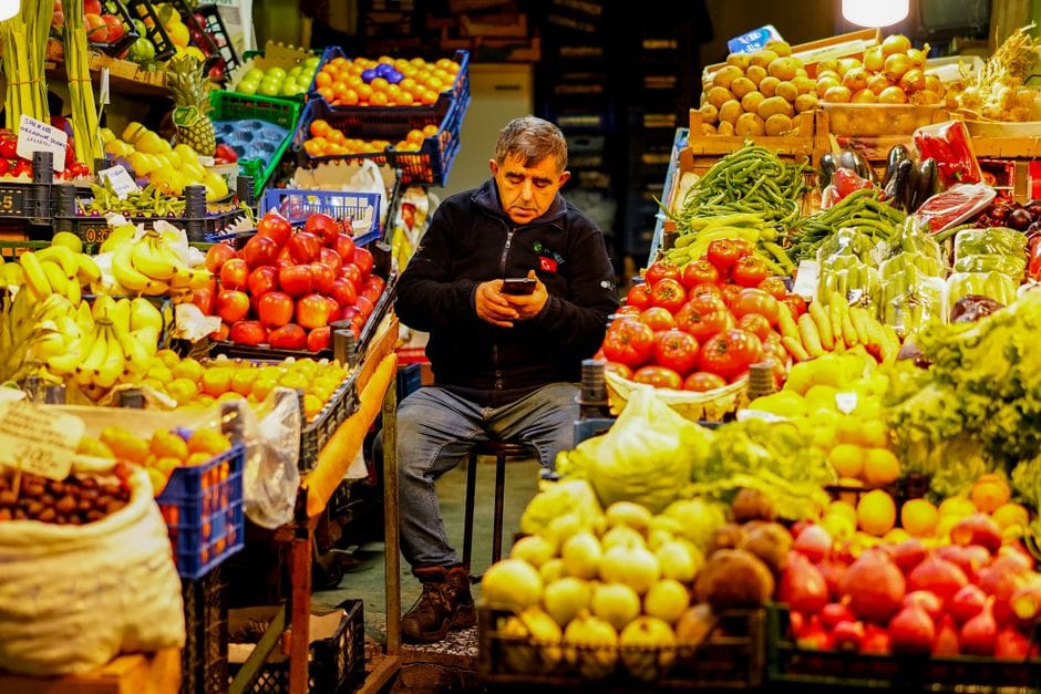 Hombre sentado en un colorido puesto de mercado, rodeado de frutas y verduras frescas.