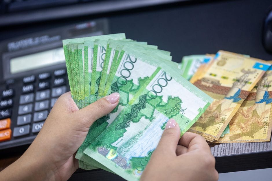 Primer plano de manos contando billetes de tenge kazajo en un escritorio con calculadora.