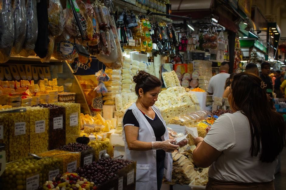 Un mercado interior abarrotado de gente con un vendedor de queso atendiendo a los clientes en medio de una variedad de alimentos gourmet.