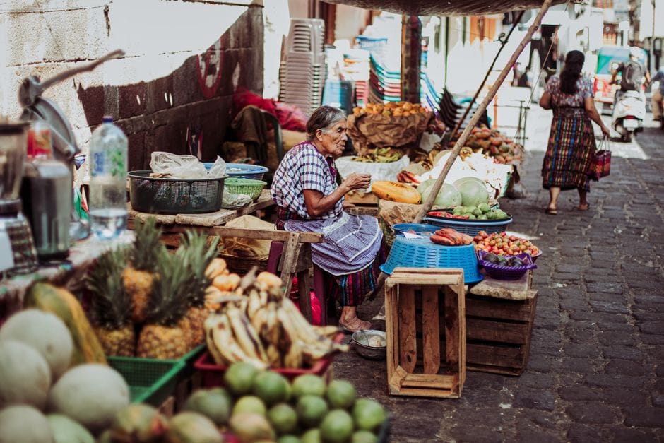 Vendedora anciana en un bullicioso mercado callejero rodeada de frutas y verduras frescas en un día soleado.