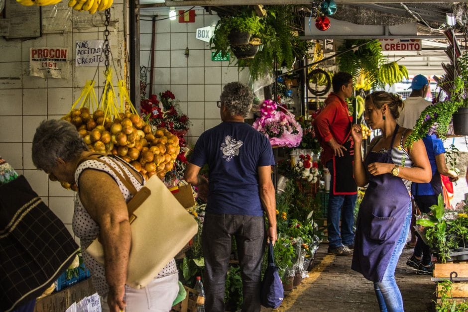 Escena vibrante de mercado interior con compradores diversos entre coloridos puestos de flores y frutas.