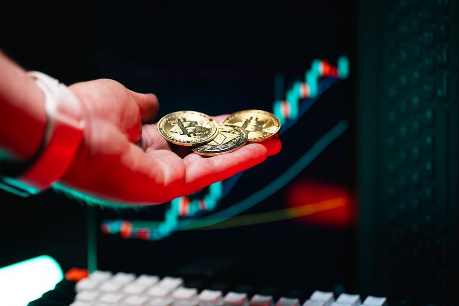 Primer plano de una mano sosteniendo monedas de criptomonedas con un gráfico comercial en el fondo.
