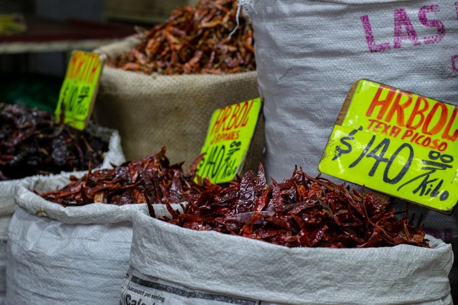Primer plano de chiles secos en sacos en un mercado, con etiquetas de precio escritas a mano en colores vivos.