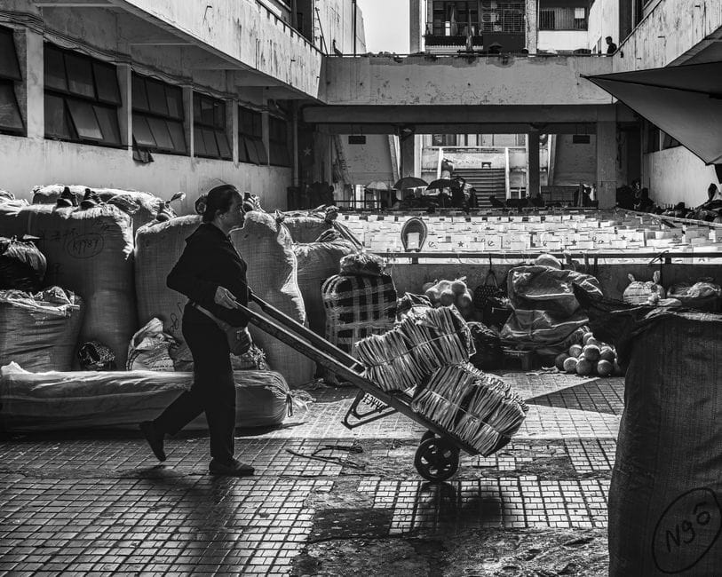 Una mujer en un mercado empuja un carrito lleno de mercancías en un entorno urbano, en blanco y negro.