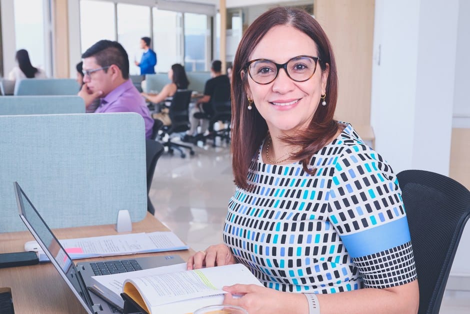 Mujer segura de sí misma en una oficina moderna, rodeada de colegas, trabajando en una computadora portátil.