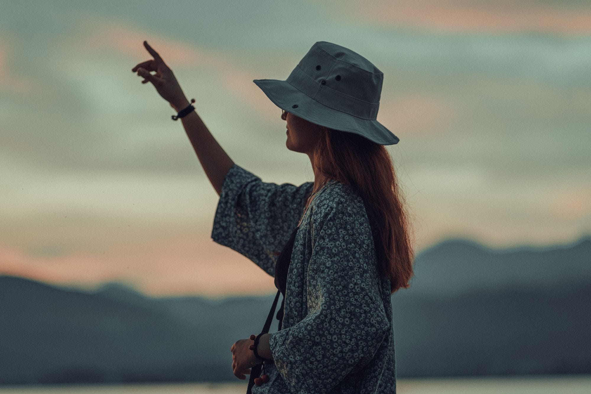 Silhouette of a woman in a bucket hat pointing towards a picturesque evening sky with mountains in the background.