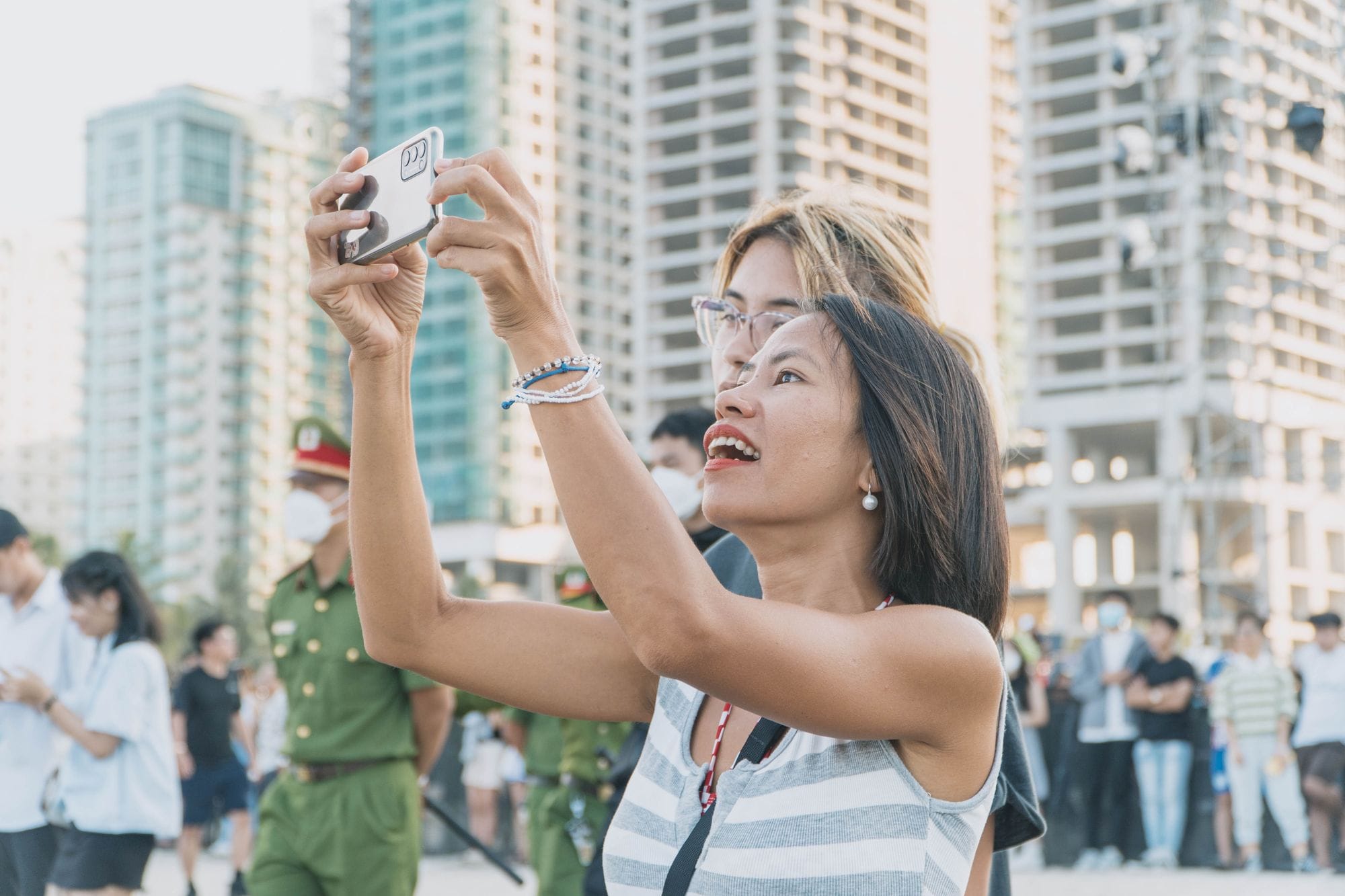An Asian woman joyfully takes a photo outdoors in a bustling urban setting. Captures a lively atmosphere.