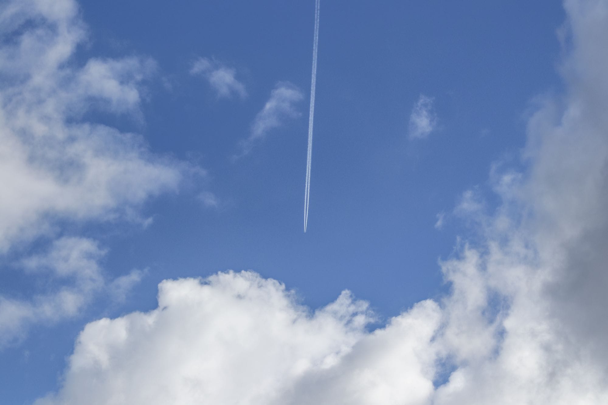 A high-altitude airplane contrail streaks across a bright blue sky with scattered white clouds.