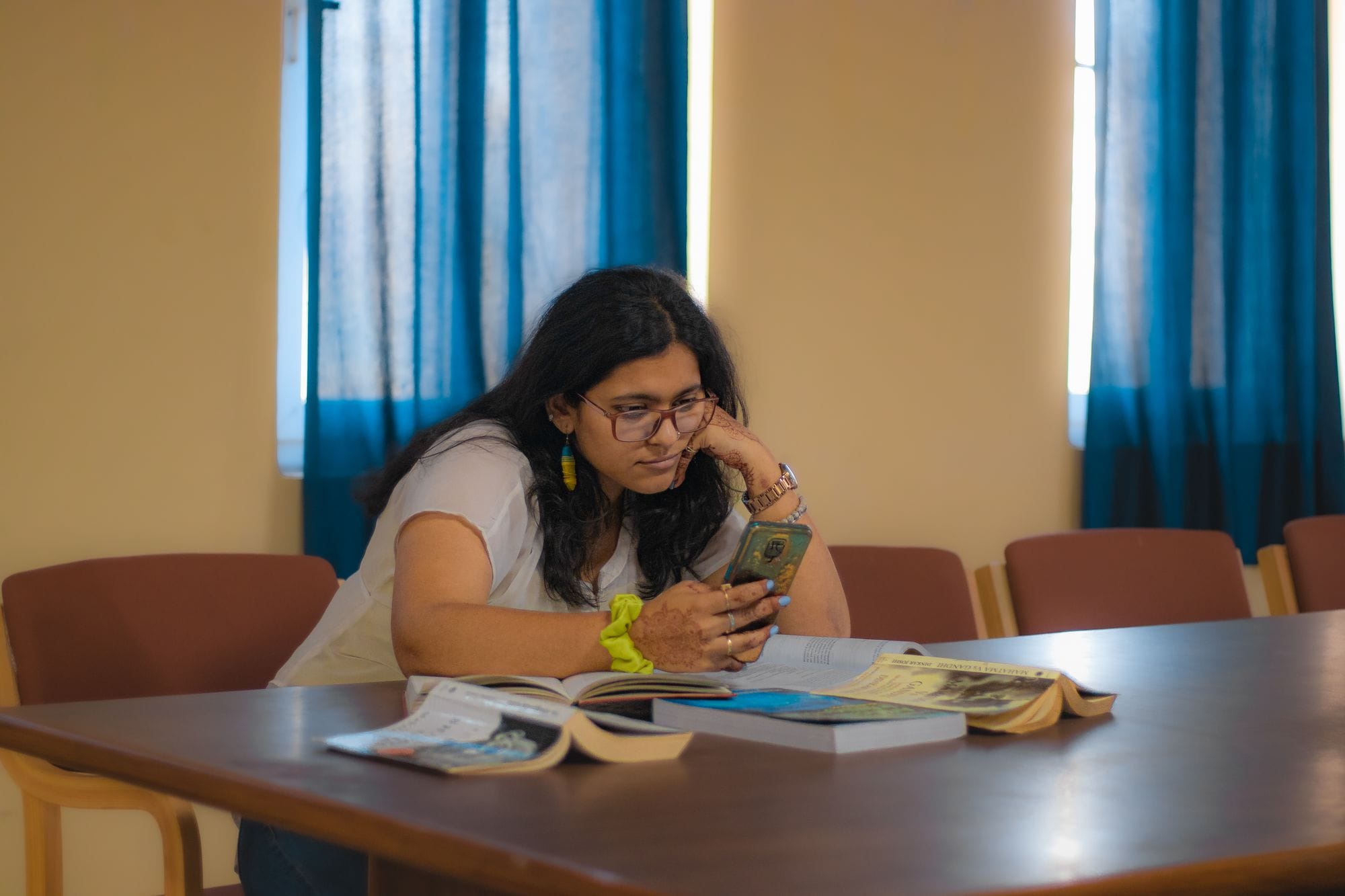 Woman in Delhi library studying with books and smartphone, wearing glasses.