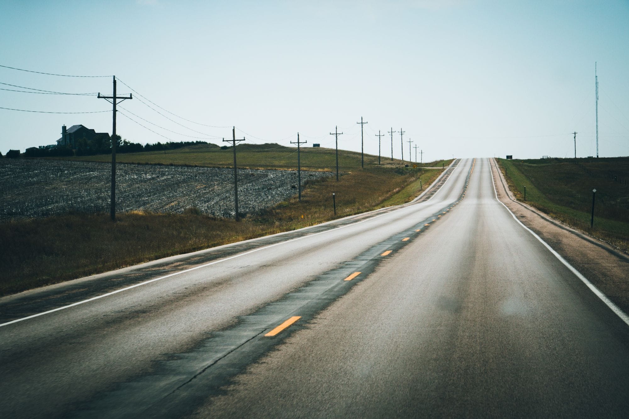 A peaceful rural highway stretching into the distance, flanked by telephone poles and open fields under a clear summer sky.