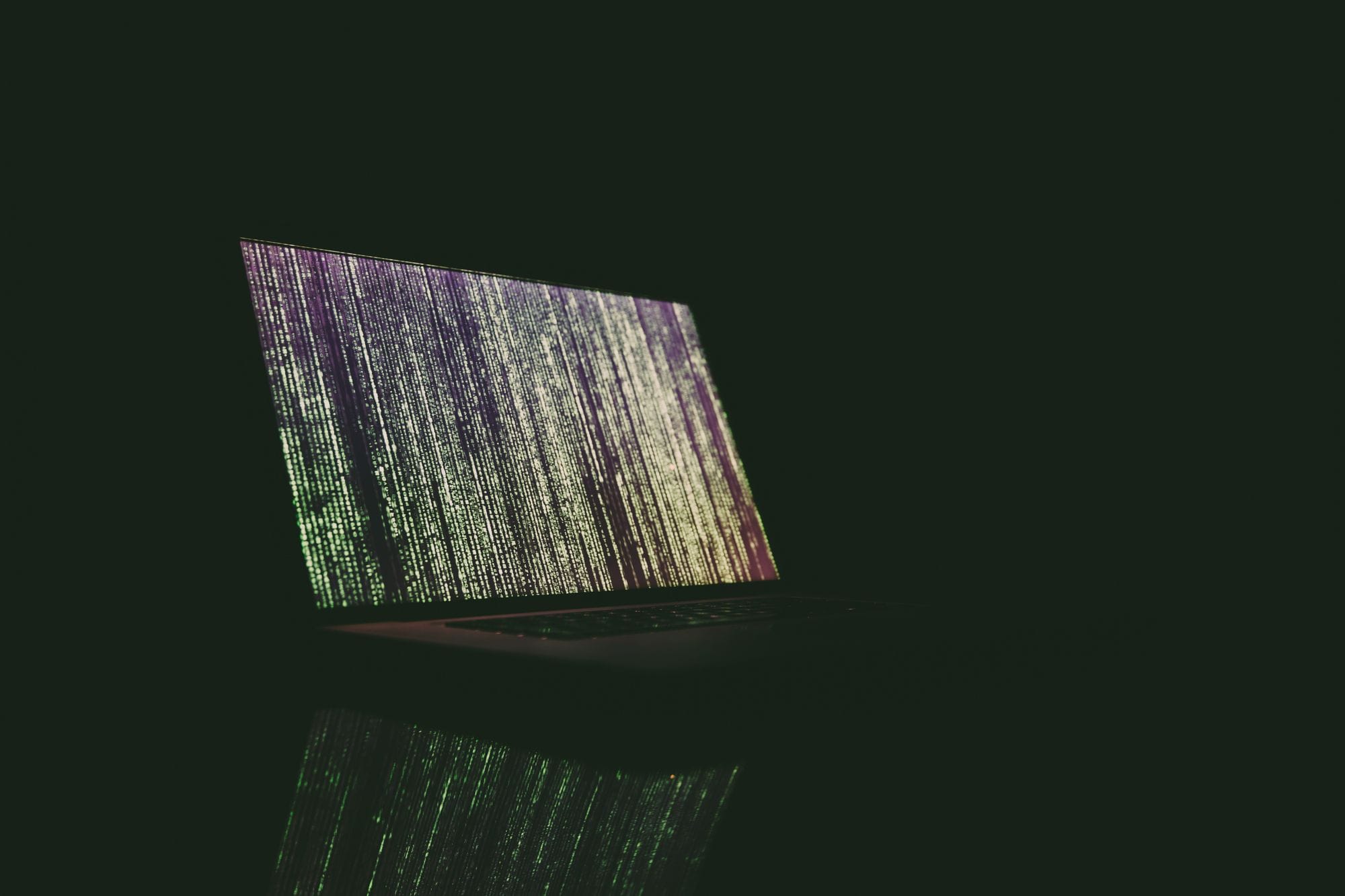 A laptop displaying green and purple code in a dark room, reflecting on a surface.