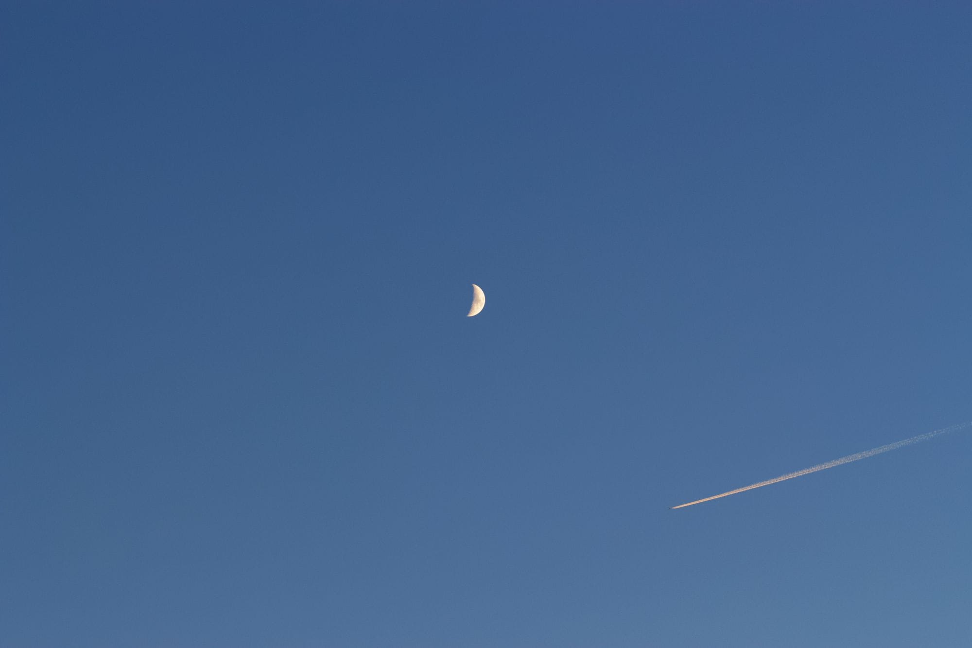 A crescent moon and airplane contrail against a clear blue sky, creating a serene contrast.