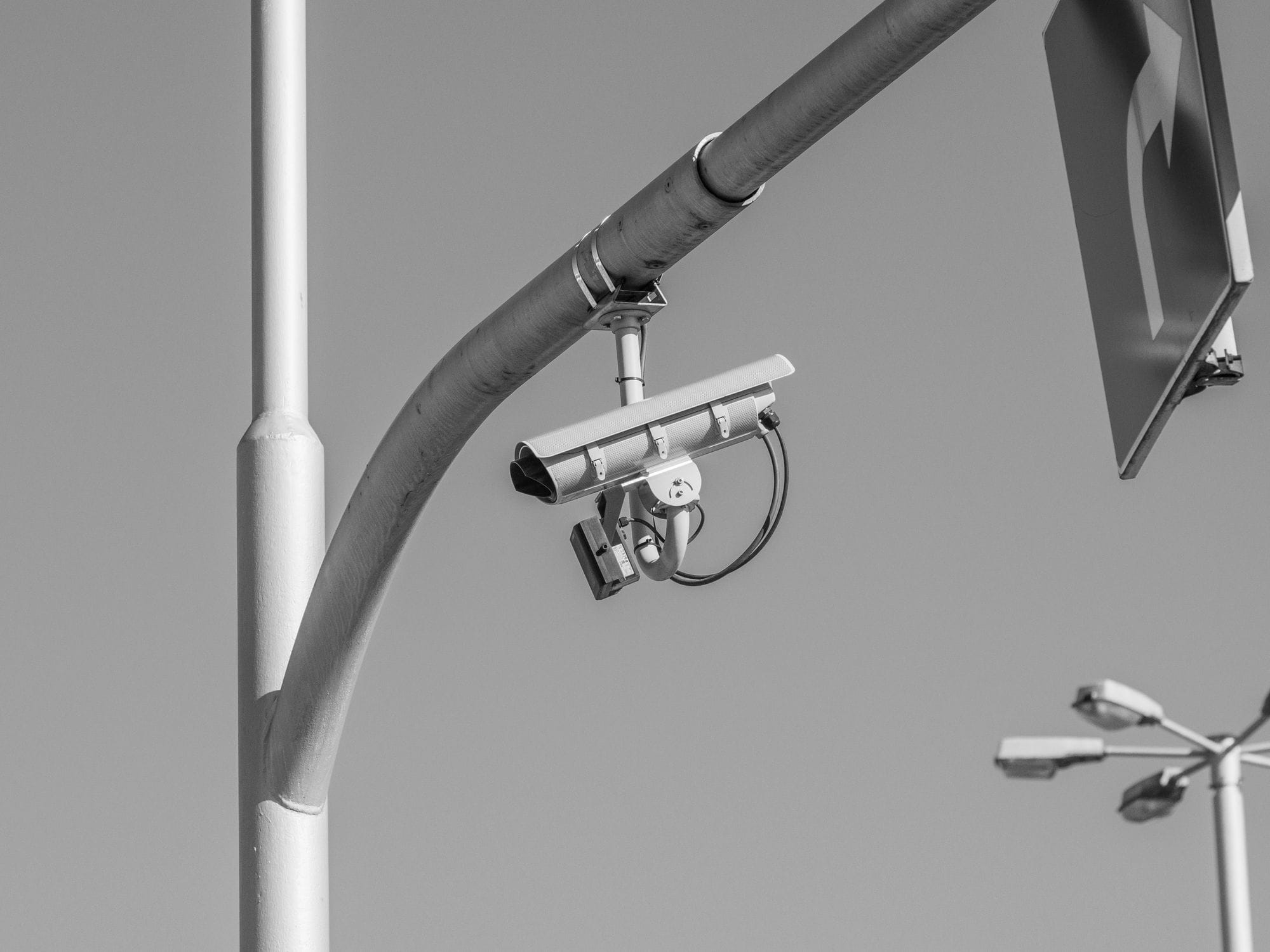 Black and white image of a surveillance camera mounted on a streetlight in an urban environment.