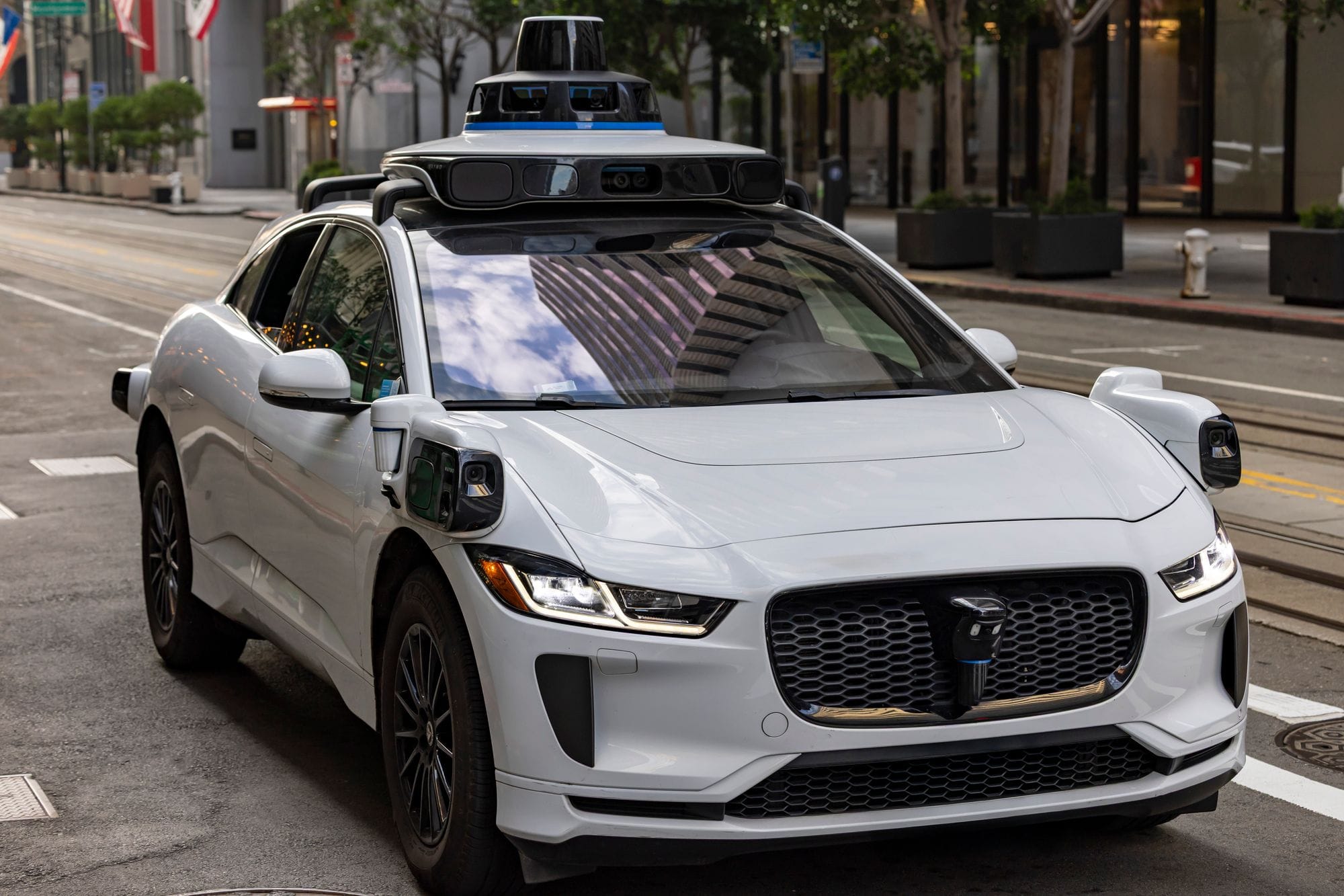 A white autonomous vehicle navigating a city street, reflecting urban architecture in daylight.