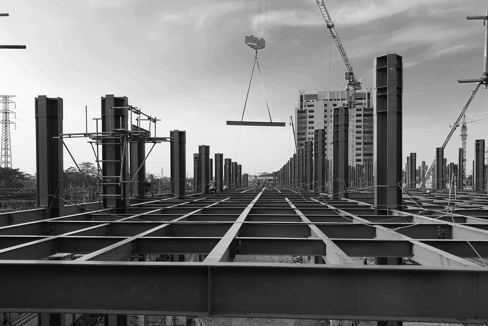 Black and white view of a modern urban construction site in Indonesia with steel beams and cranes against a cloudy sky.