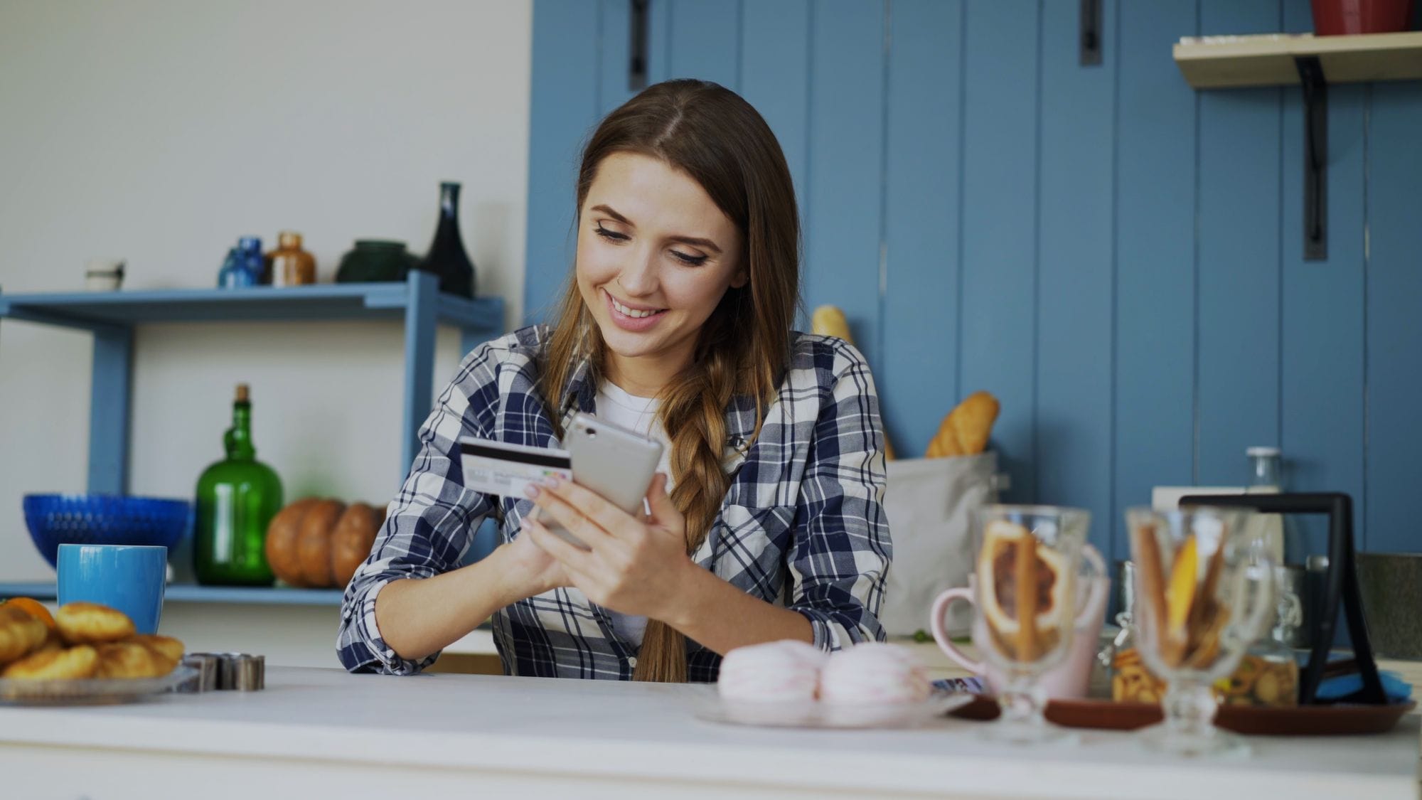 Smiling woman using smartphone and credit card for online shopping in a cozy indoor kitchen.