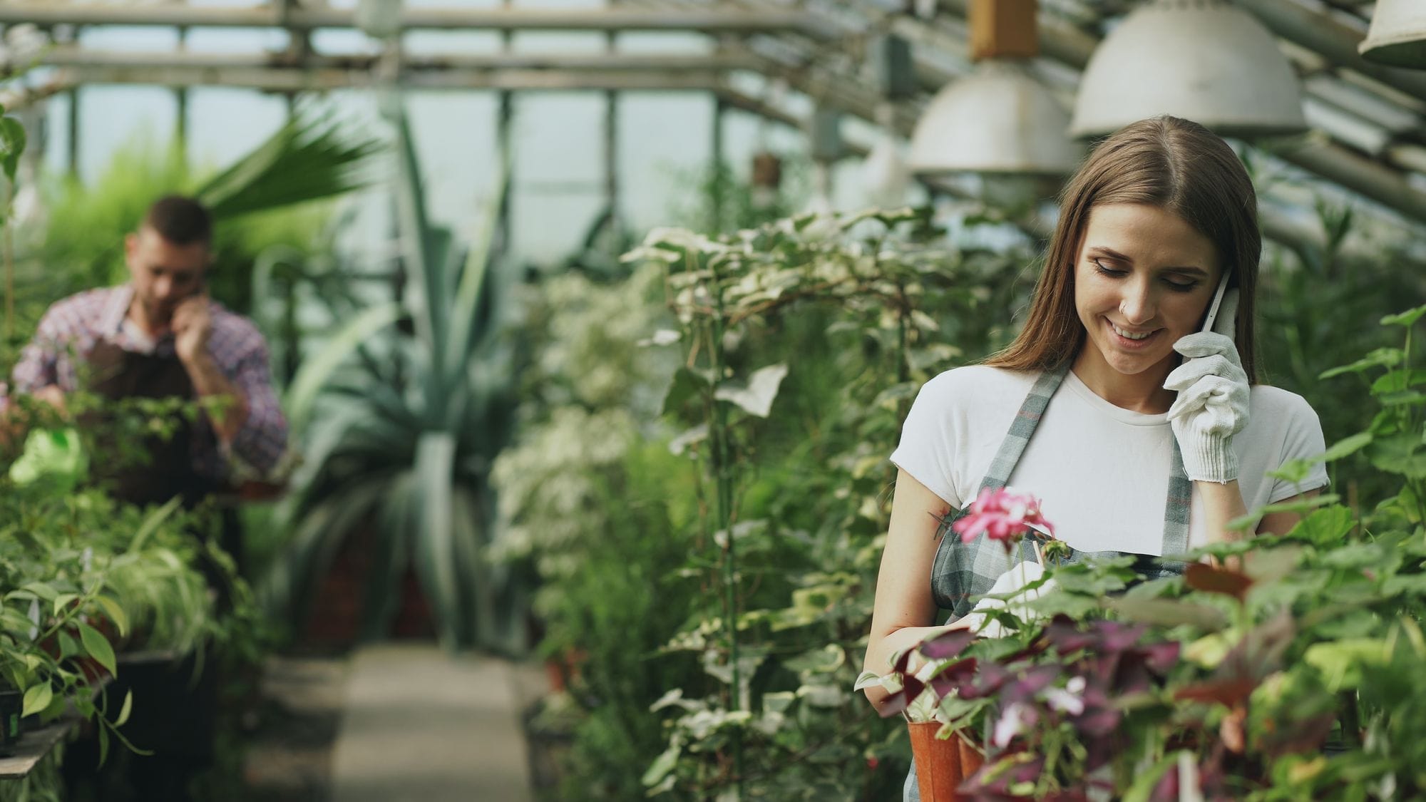 Woman gardener with white gloves talks on phone amidst lush plants in a greenhouse.