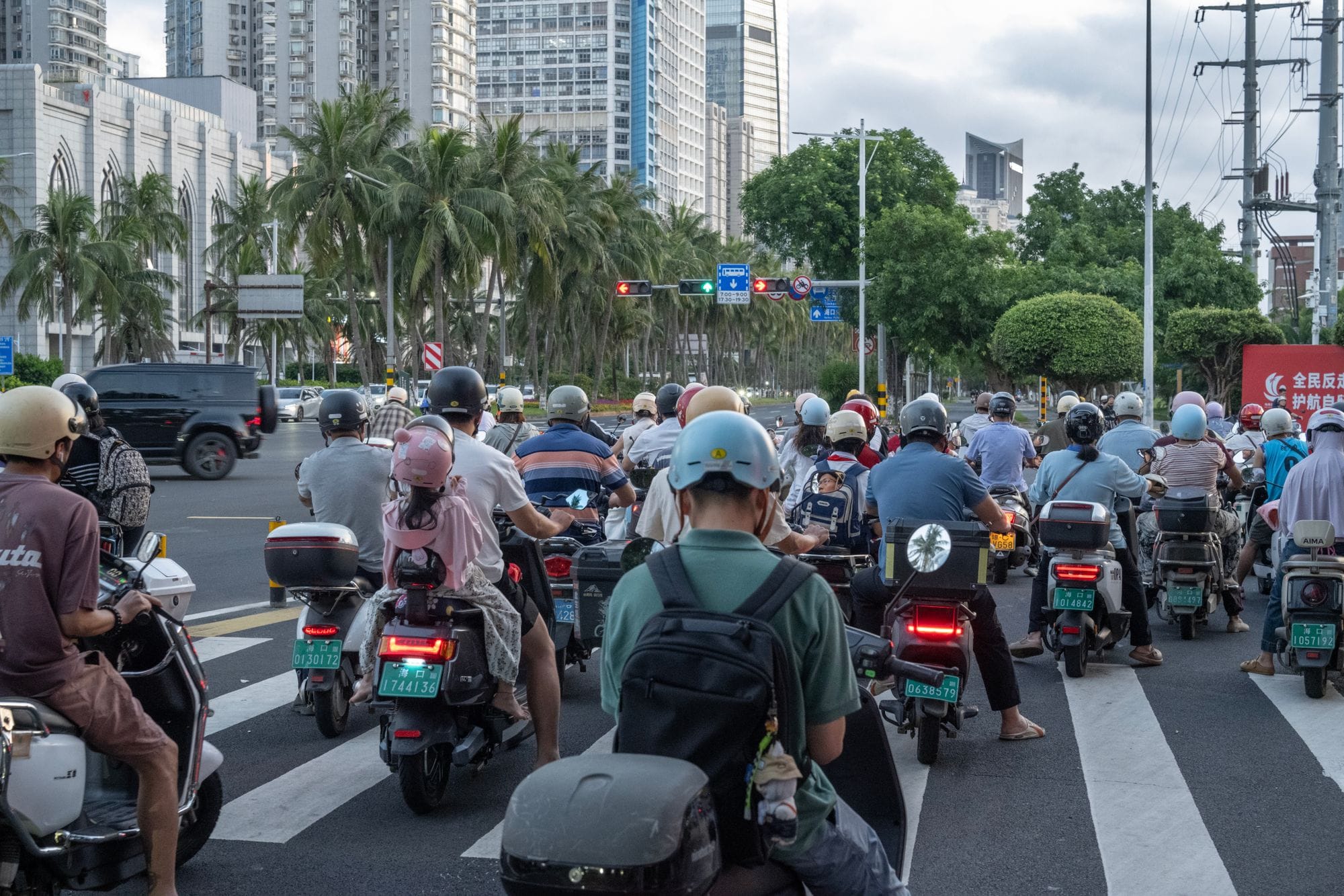 A crowded city intersection with motorcyclists stopped at a traffic light against a backdrop of modern buildings.