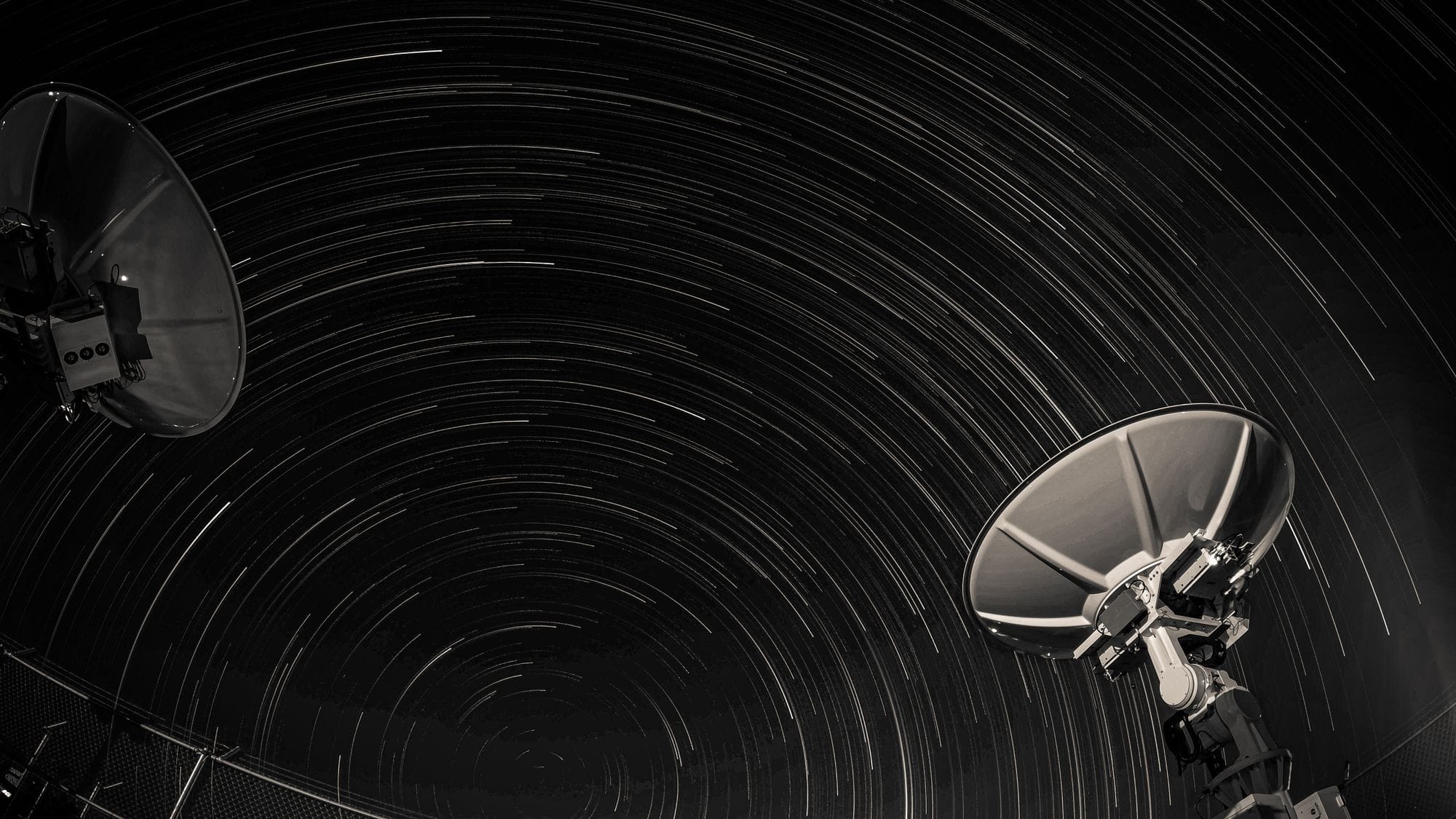 Long exposure of star trails creating circular patterns over two satellite dishes against a night sky.