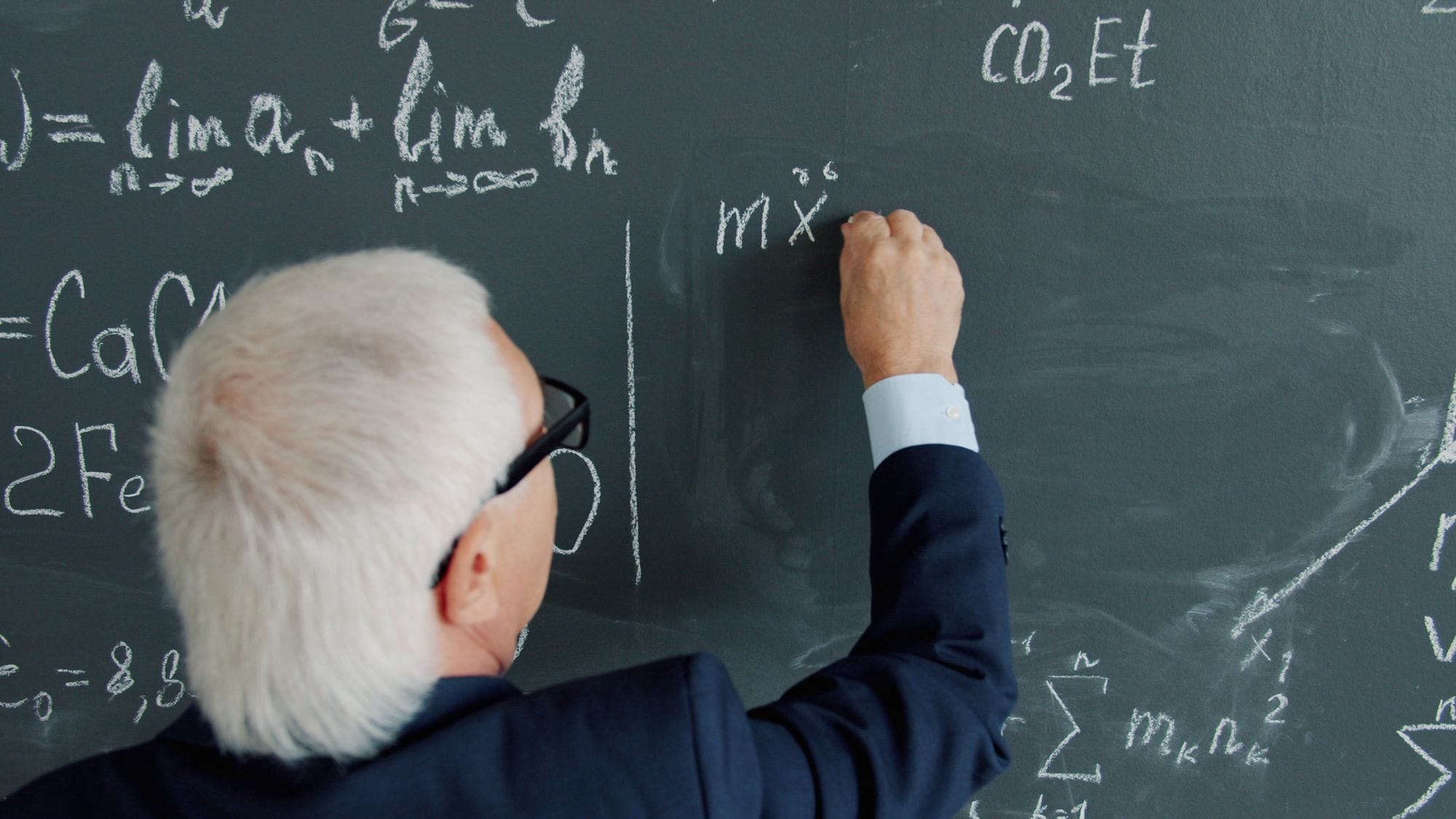 Senior male professor writing complex equations on a chalkboard.
