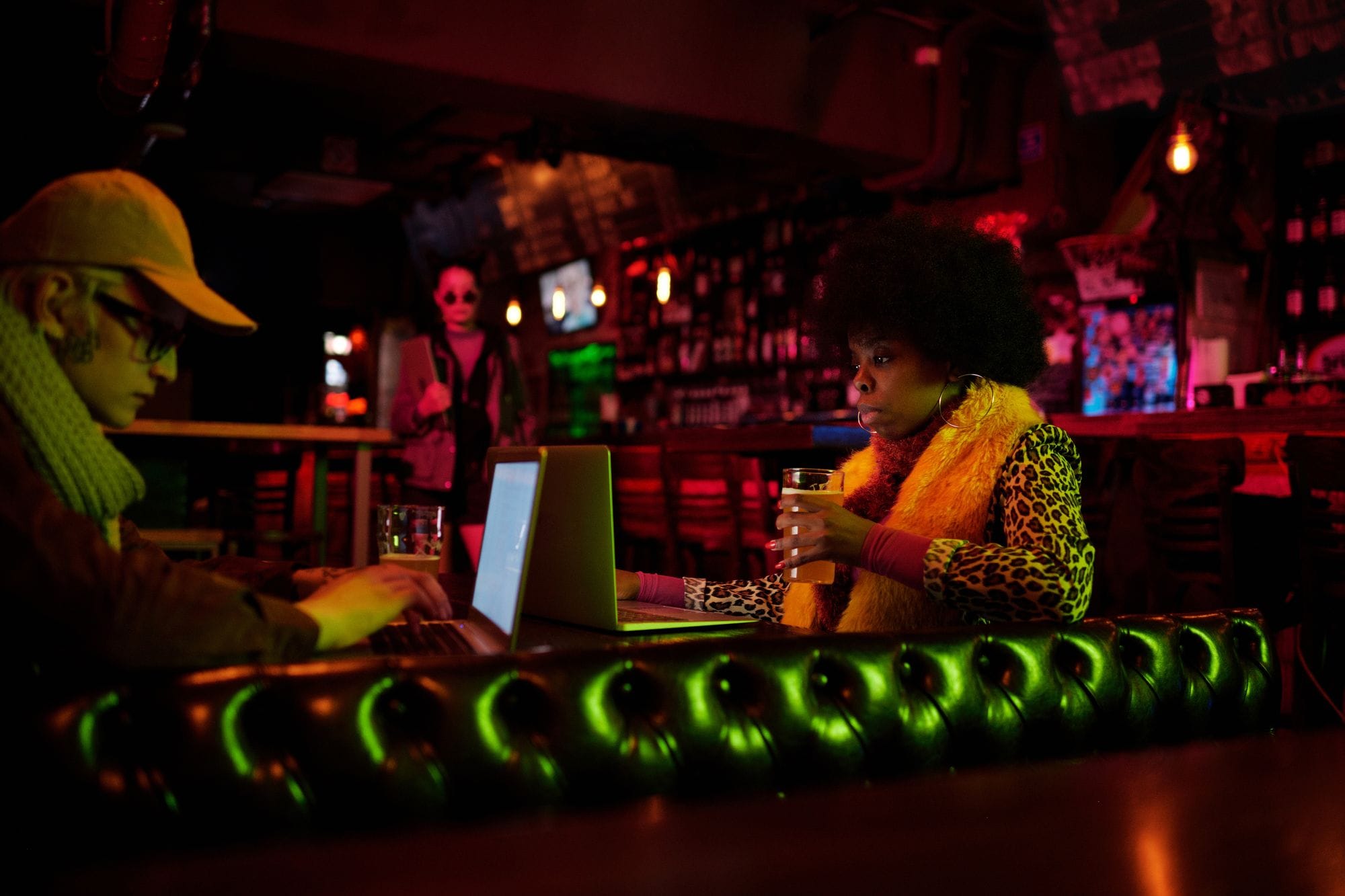Two adults working on laptops in a dimly lit bar setting, one holding a drink.