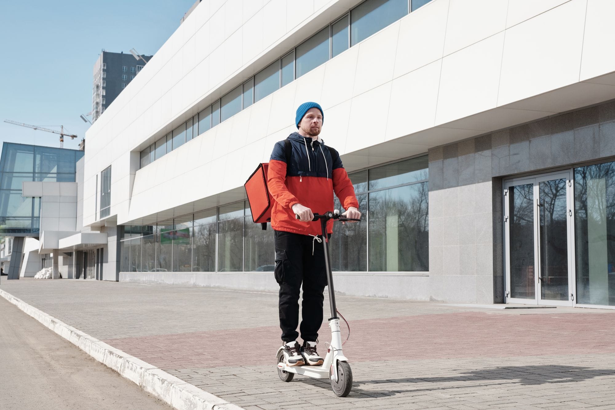 A male courier on an electric scooter delivering food outside a modern building.