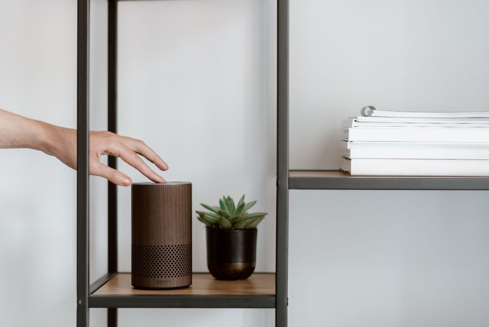 A hand interacts with a smart speaker on a minimalist shelf, featuring a plant and stacked books.