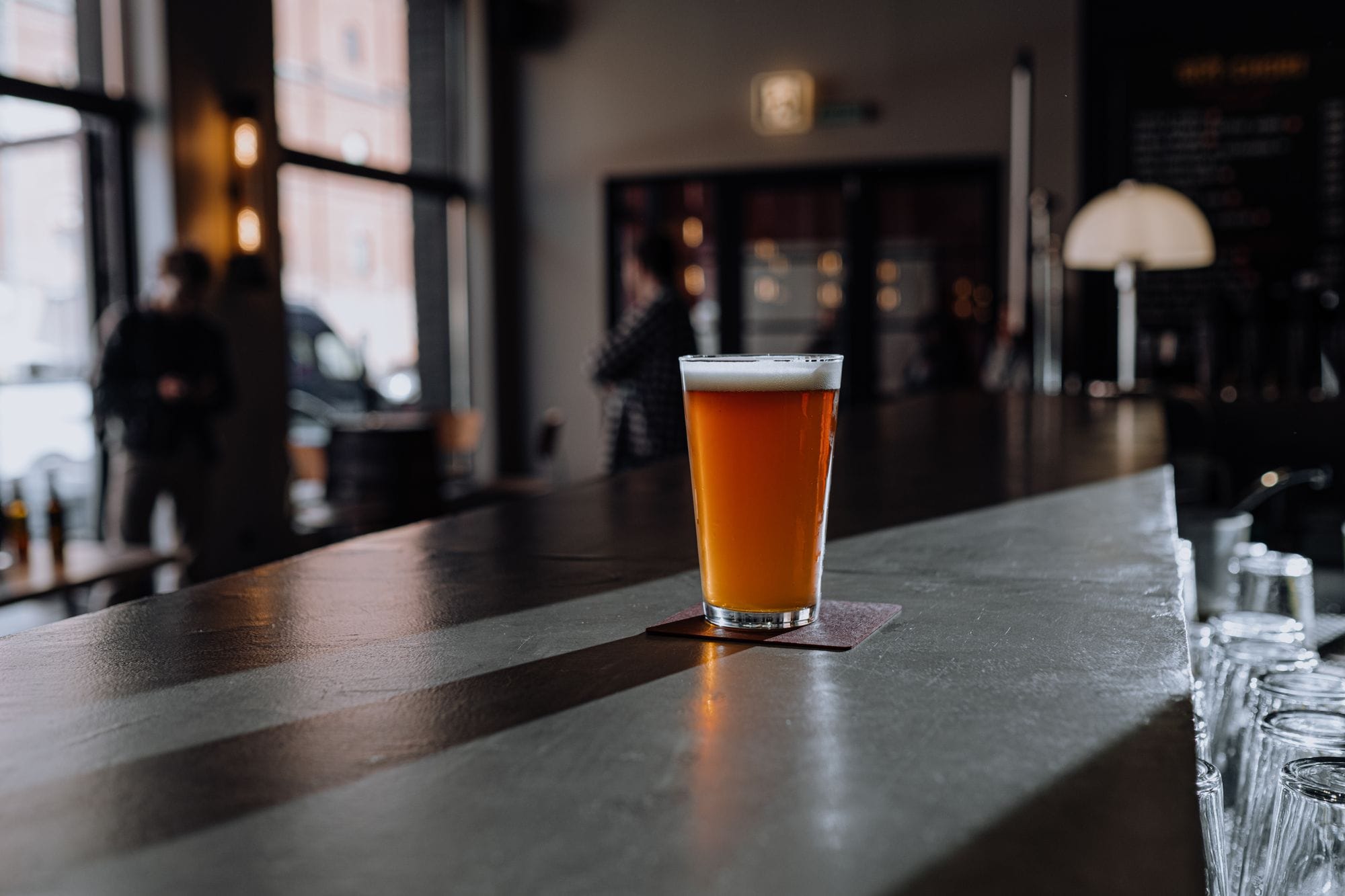 A glass of craft beer with foam sits on a bar counter in a dimly lit modern bar.