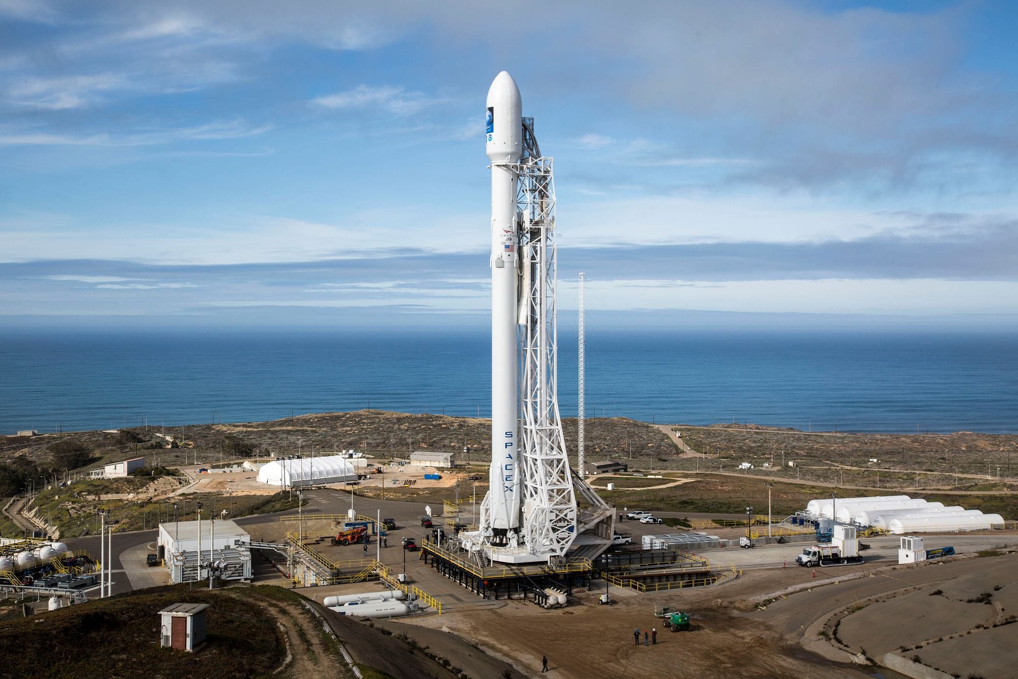 A SpaceX rocket stands on a launch pad overlooking the ocean under a clear sky.