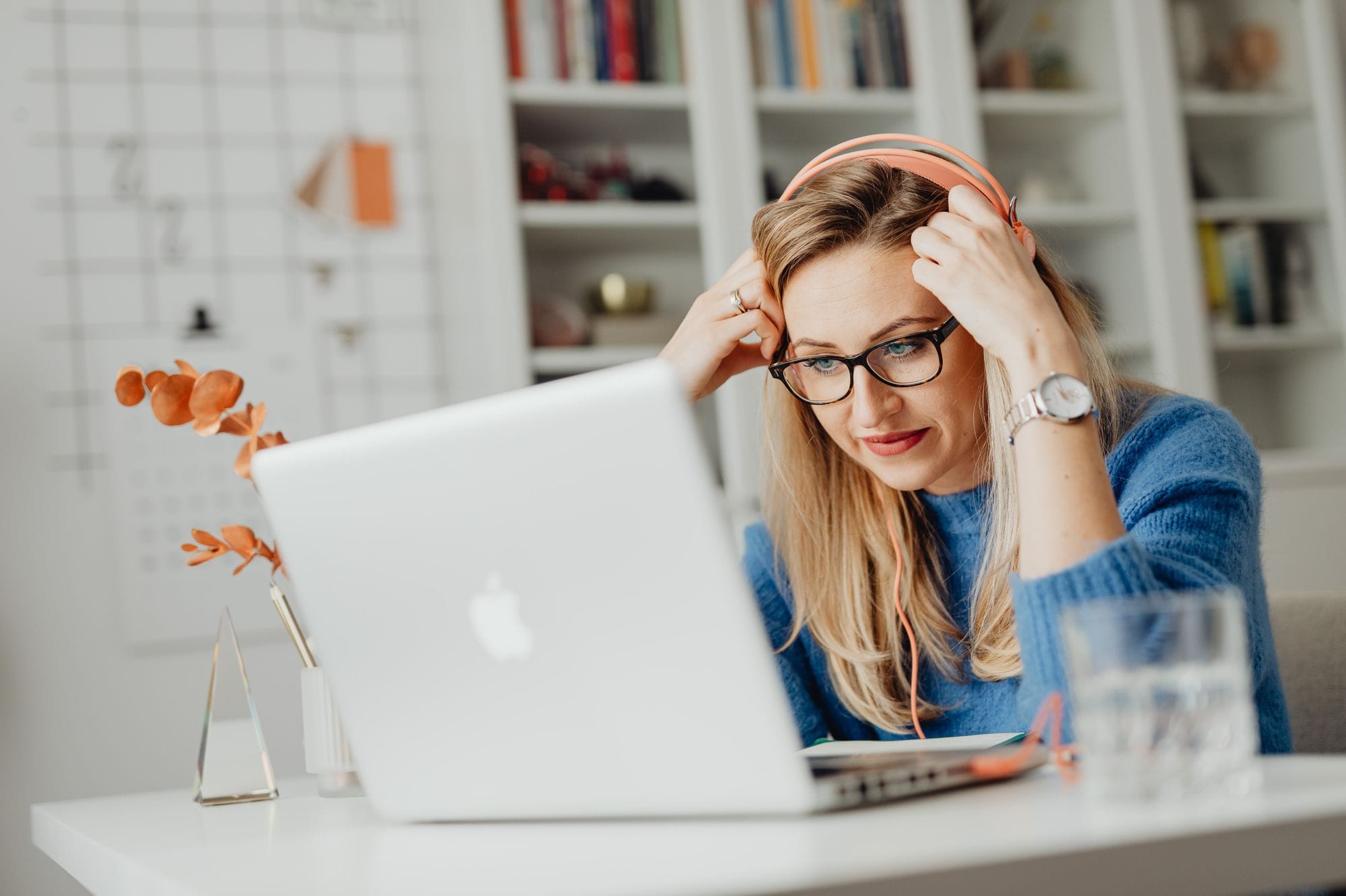 Blond woman wearing eyeglasses and headphones, working on a laptop indoors at a desk.