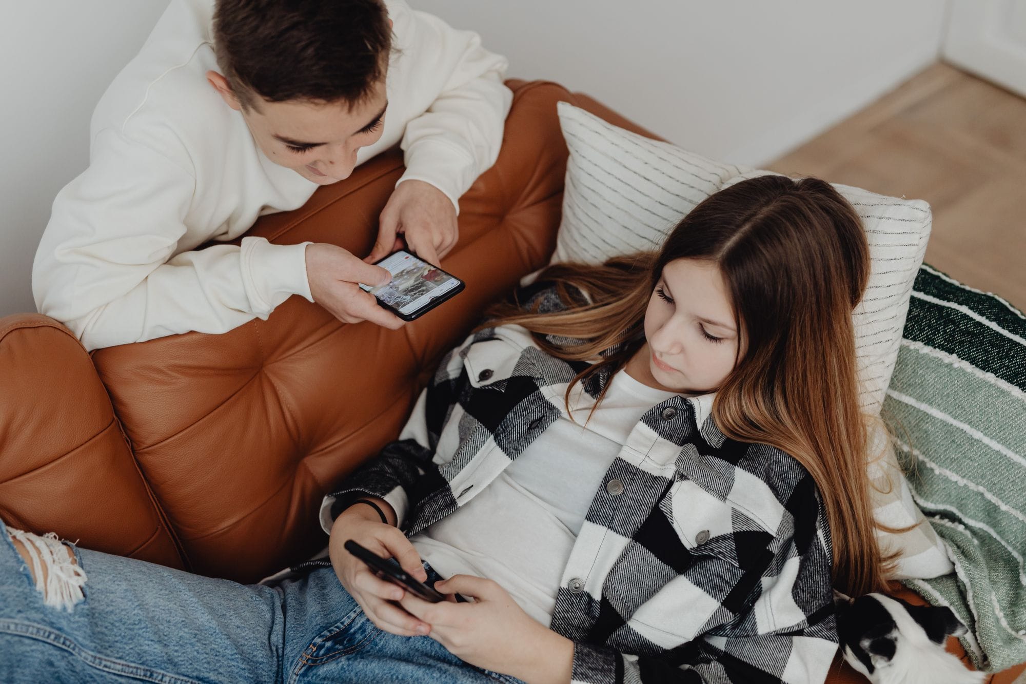 Two teenagers using smartphones while relaxing on a brown leather sofa indoors.