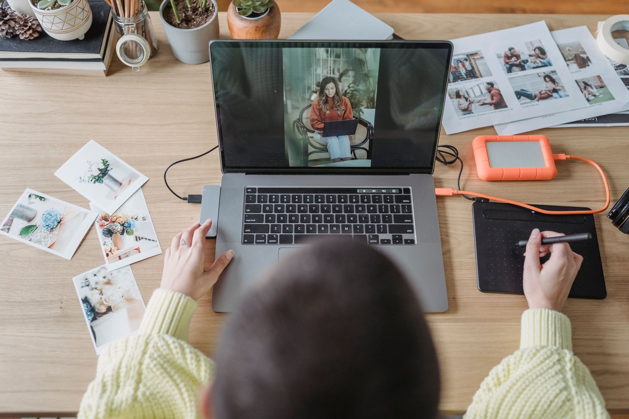 From above of crop faceless female photographer drawing on graphic tablet while editing photos on laptop at table with printed photos