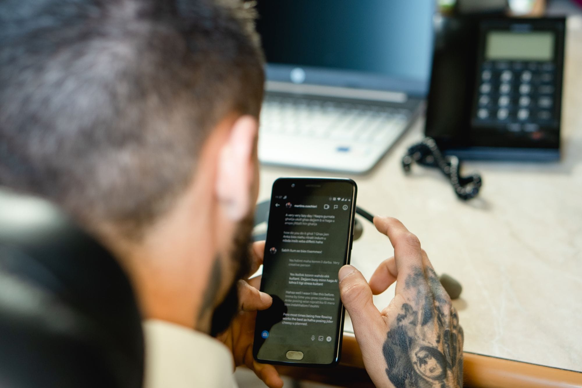 A tattooed man uses a smartphone at a desk with a laptop and a phone, symbolizing modern communication.