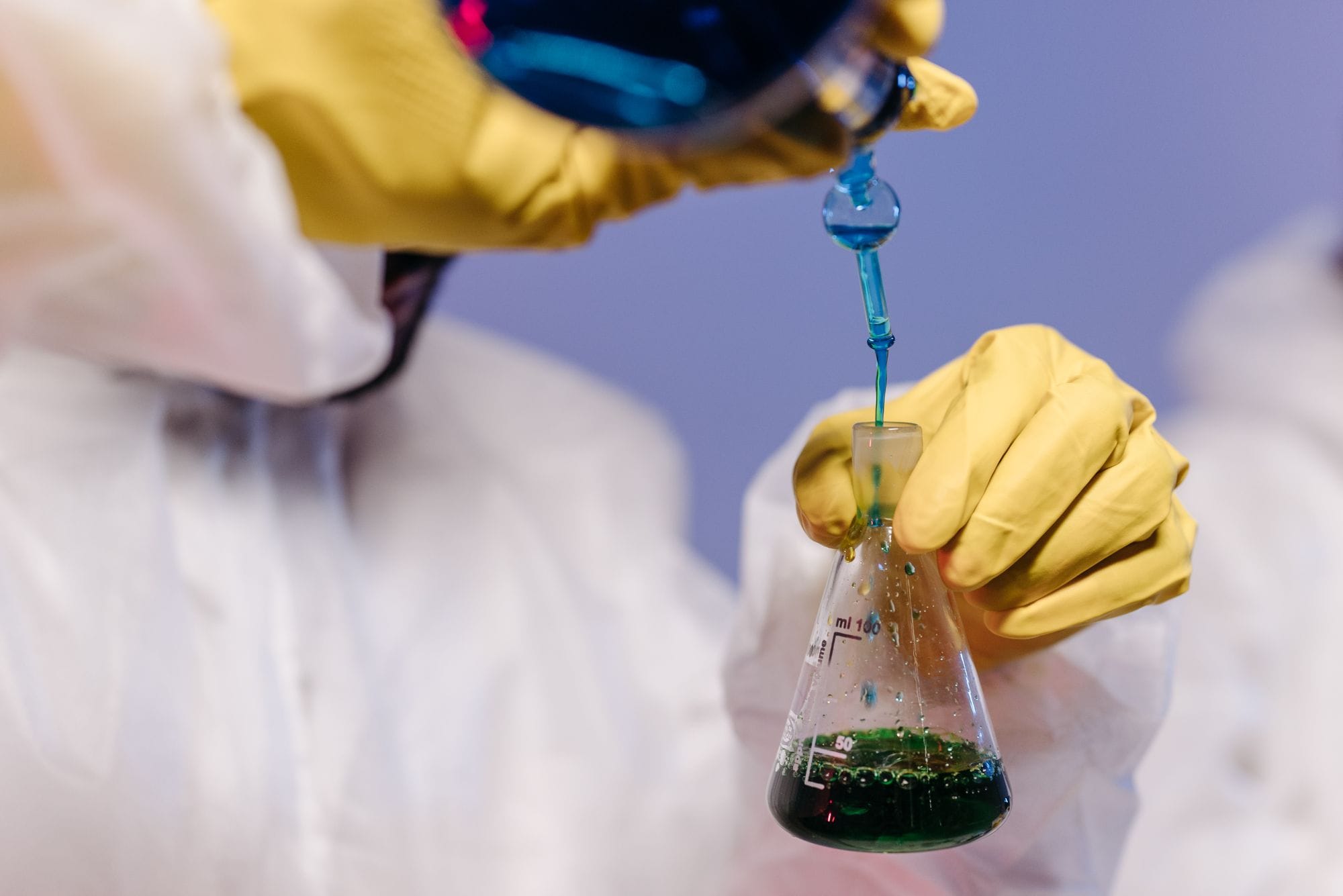 Scientist wearing gloves and lab coat mixing colored liquids in a flask.