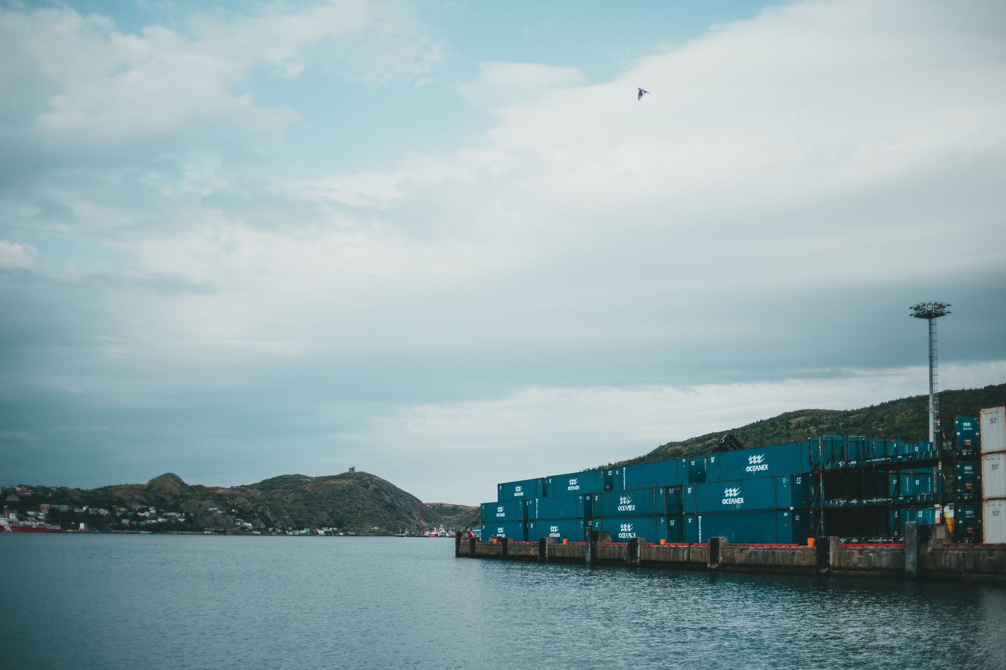 Blue shipping containers by the sea with hills under an overcast sky.
