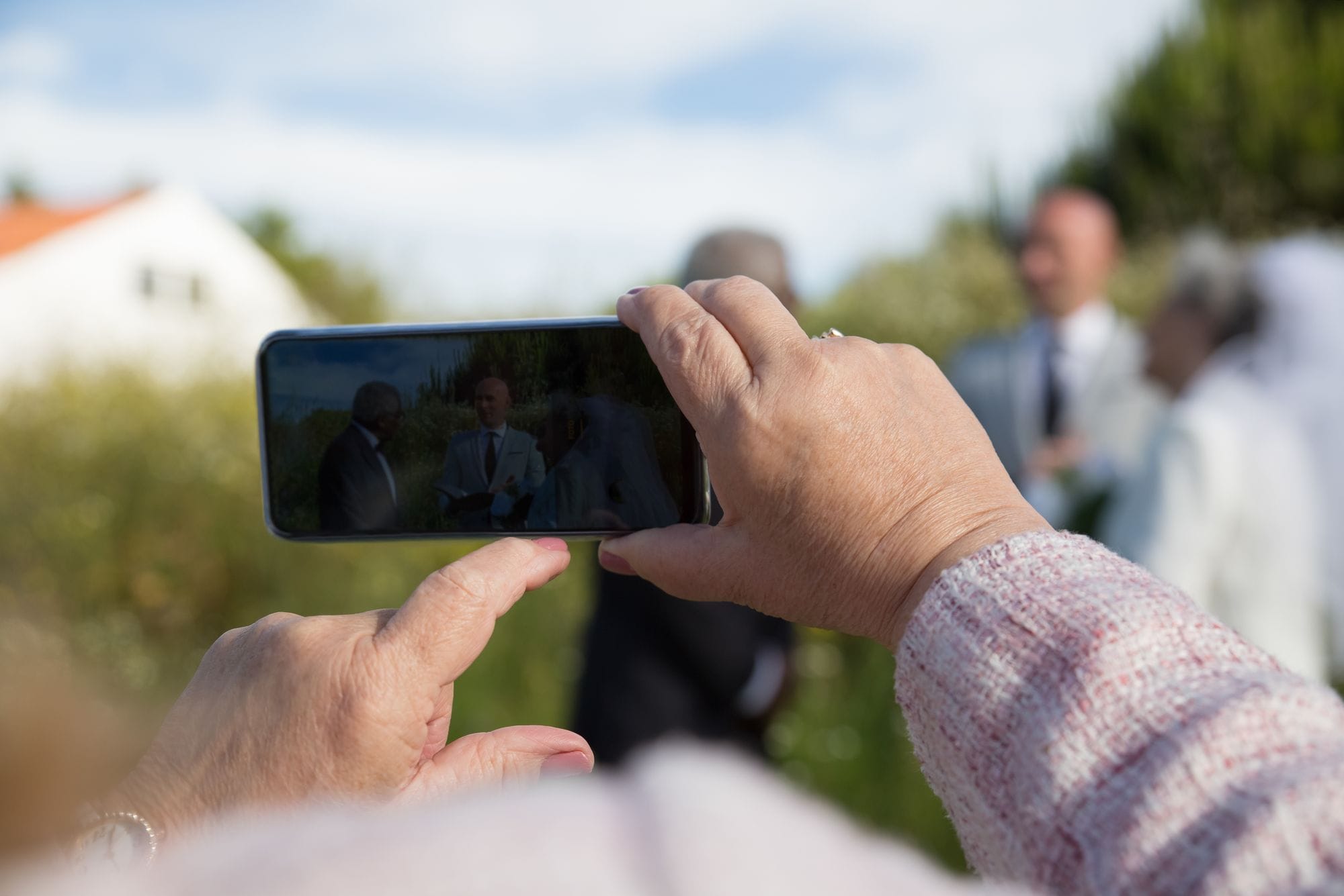 Close-up of hands using a smartphone to capture a wedding outdoors.