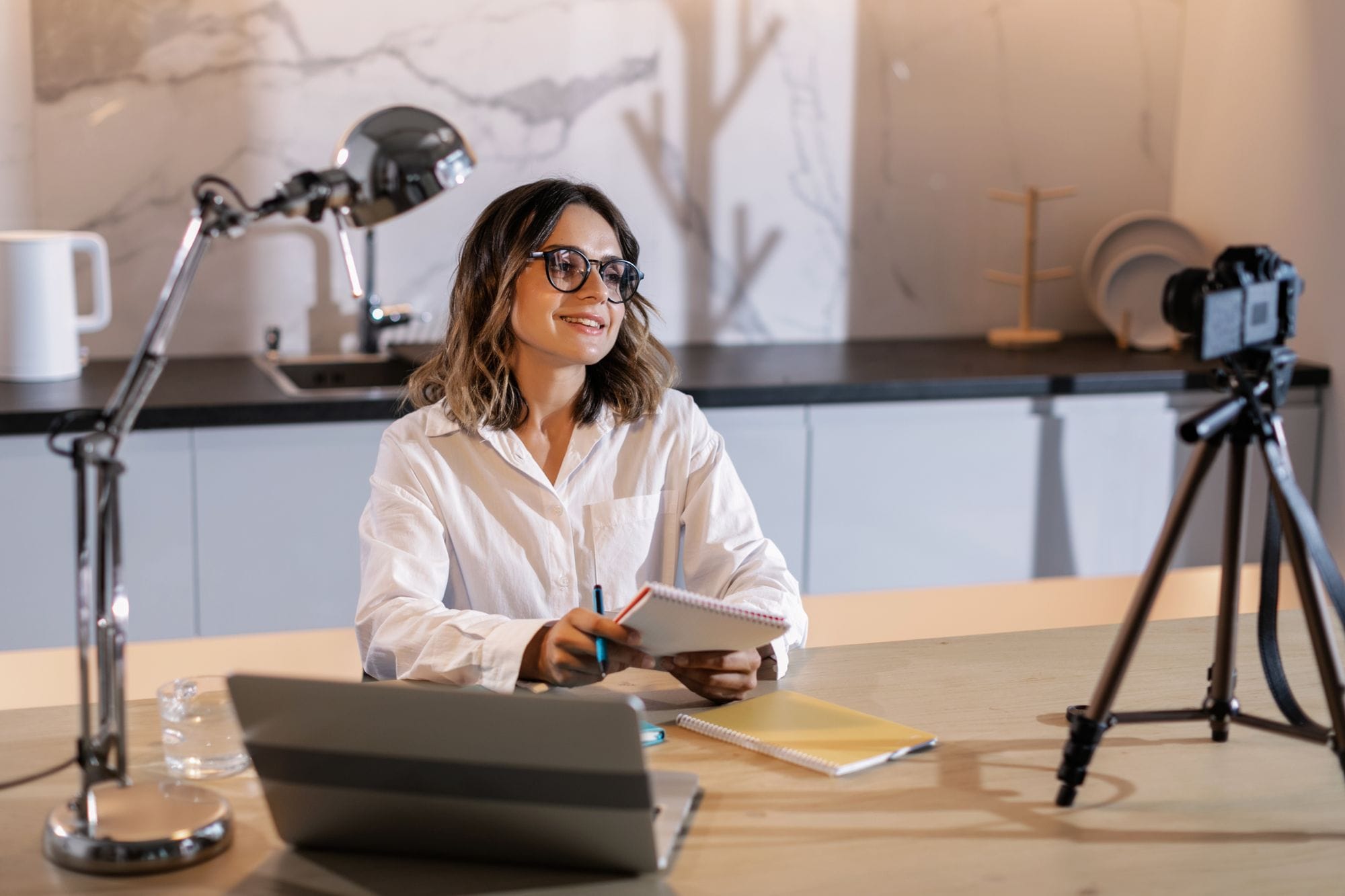 Young woman in glasses working indoors with laptop and camera, smiling while taking notes.