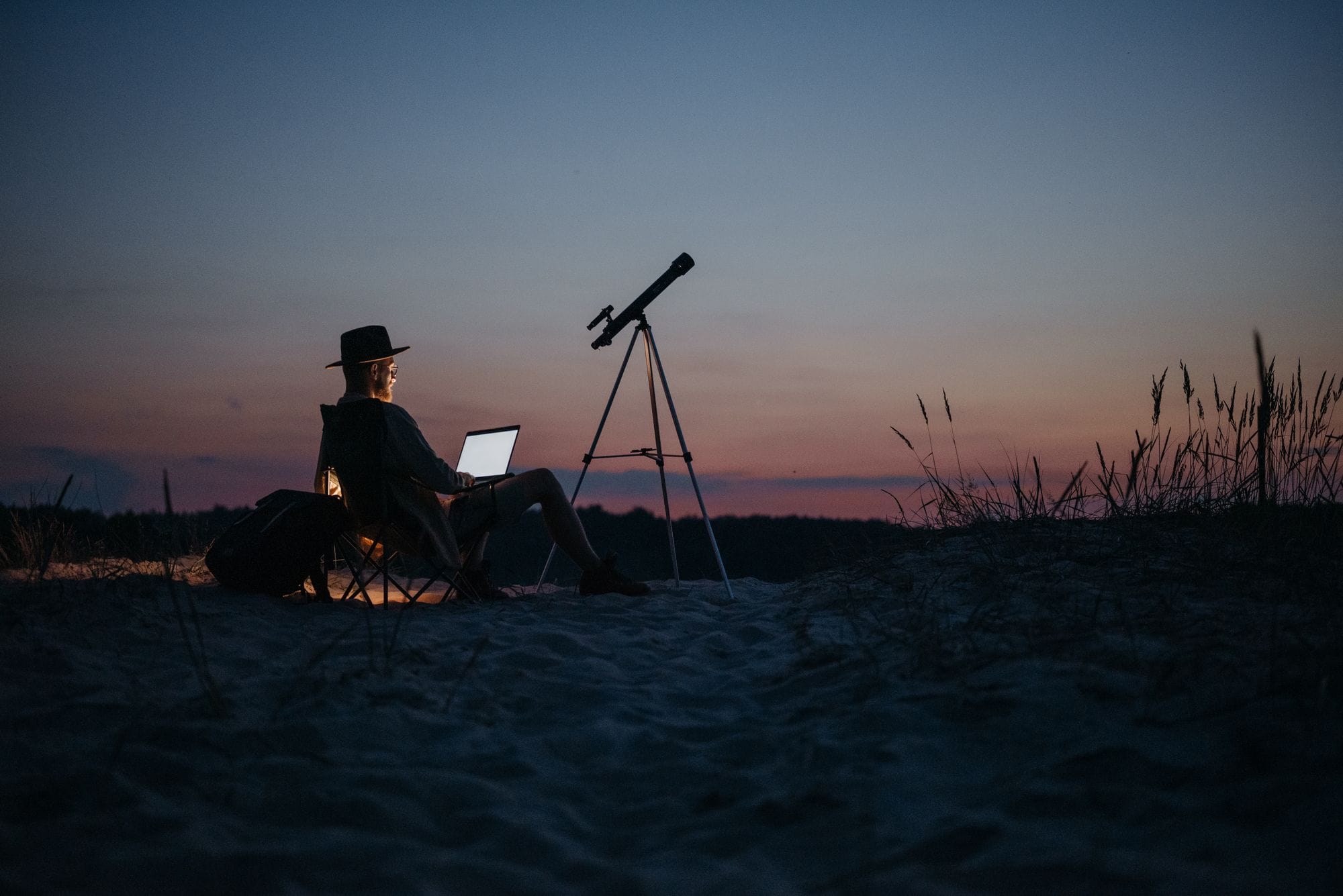 Person using a laptop and telescope at twilight, combining work and stargazing in a serene outdoor setting.