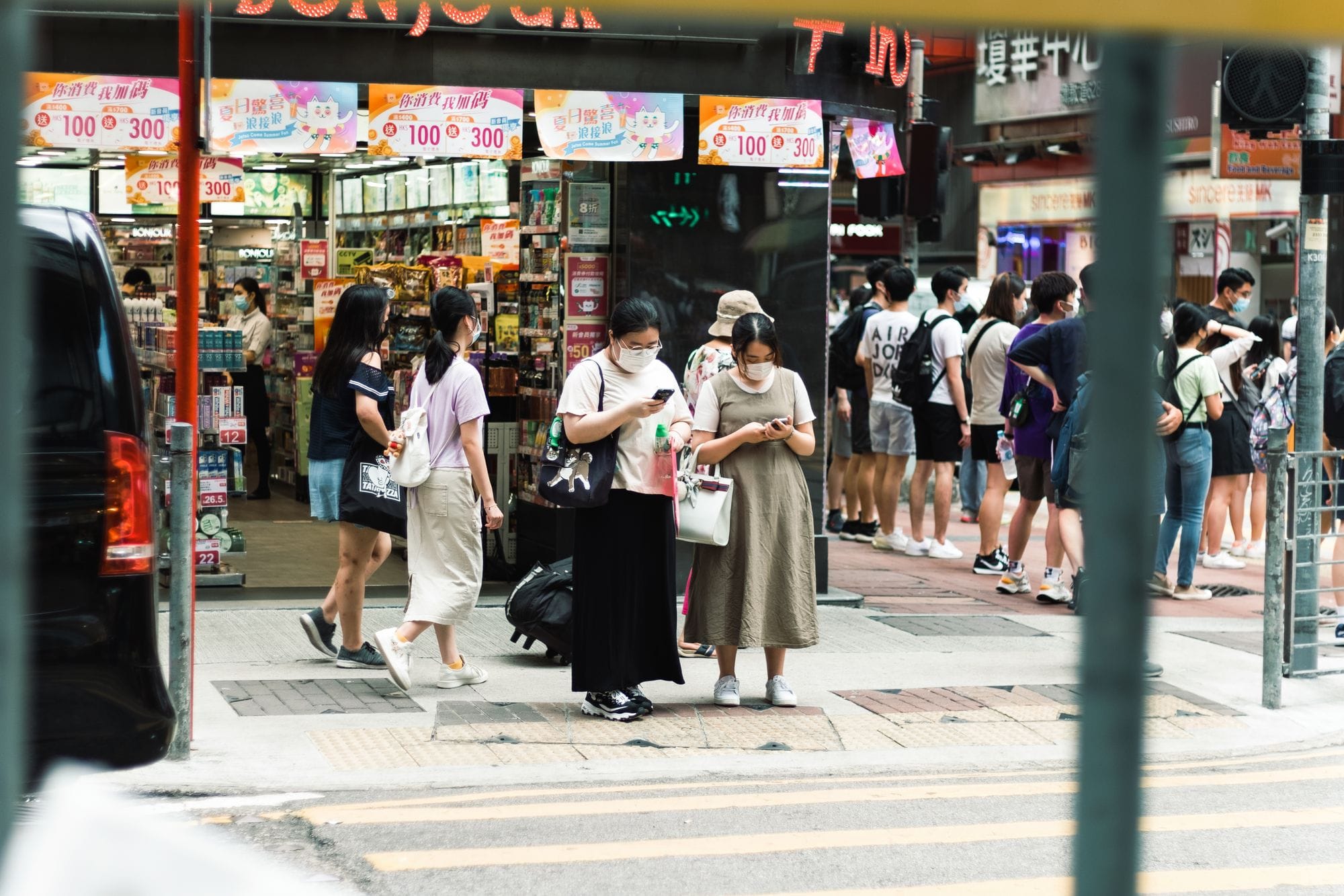 People wearing masks on a busy street near a store entrance, engaging with smartphones.