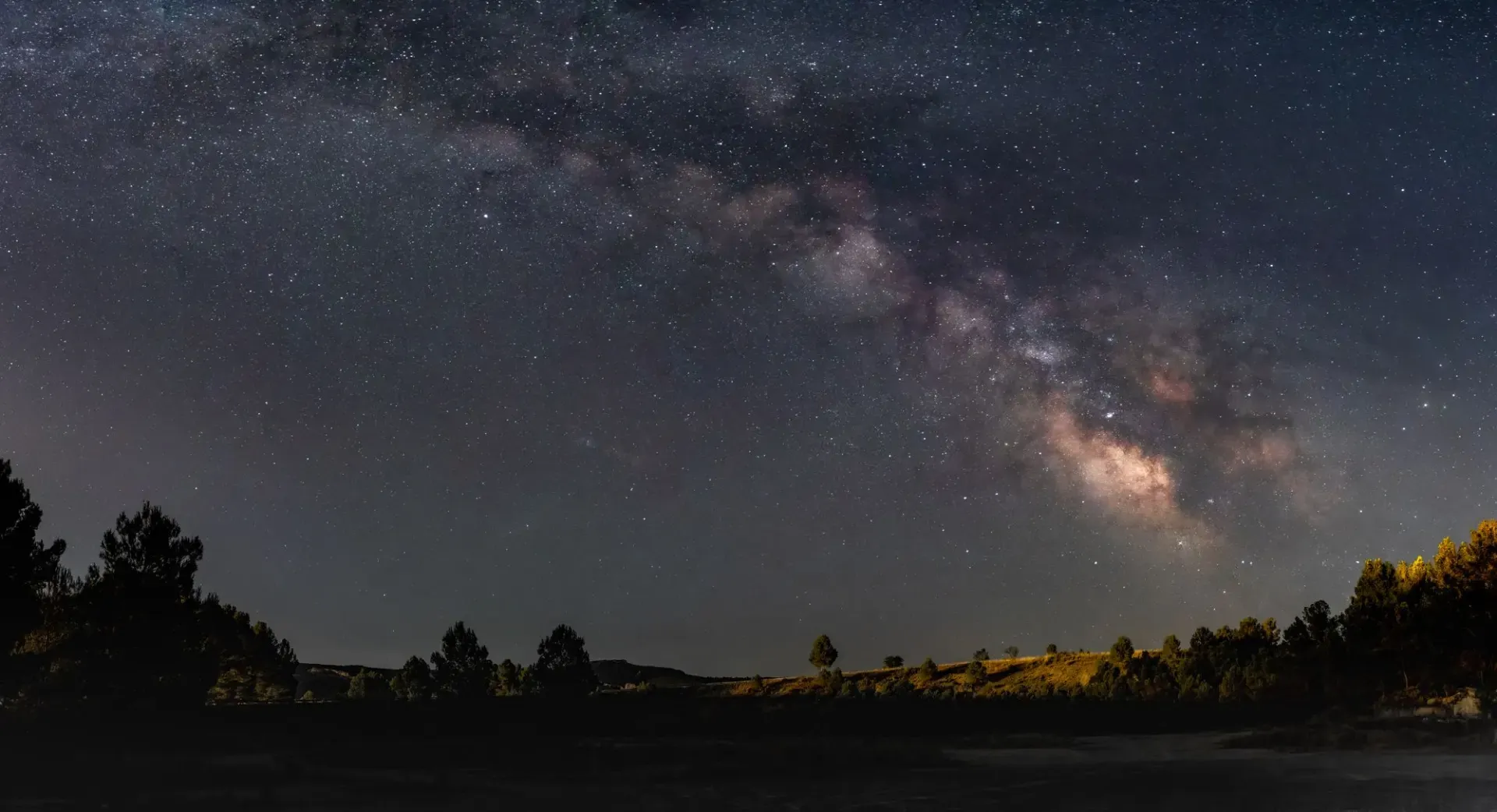 A breathtaking view of the Milky Way galaxy arching over a peaceful forest landscape at night.