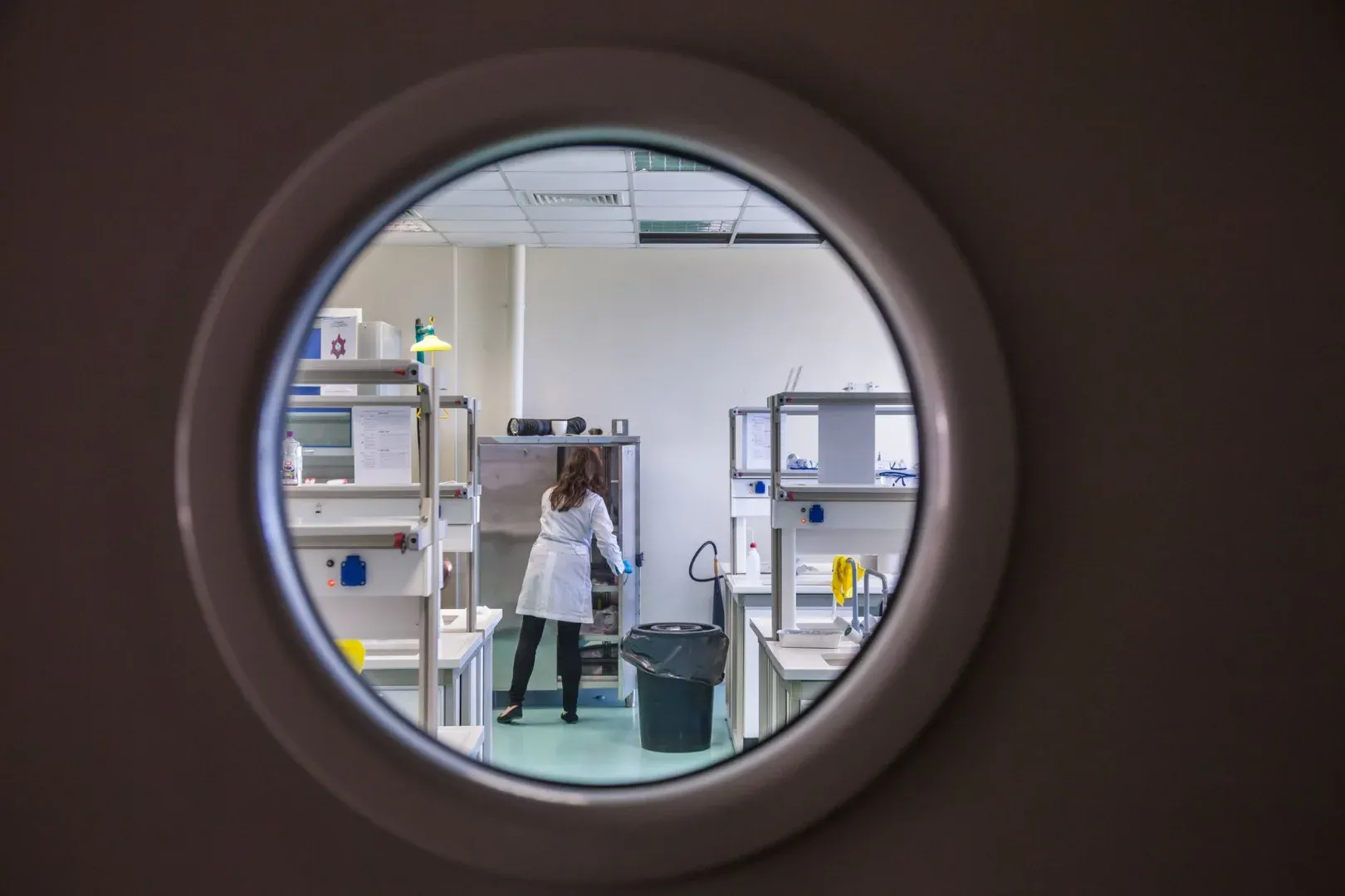 A female scientist in a lab coat working inside a state-of-the-art laboratory through a circular window view.