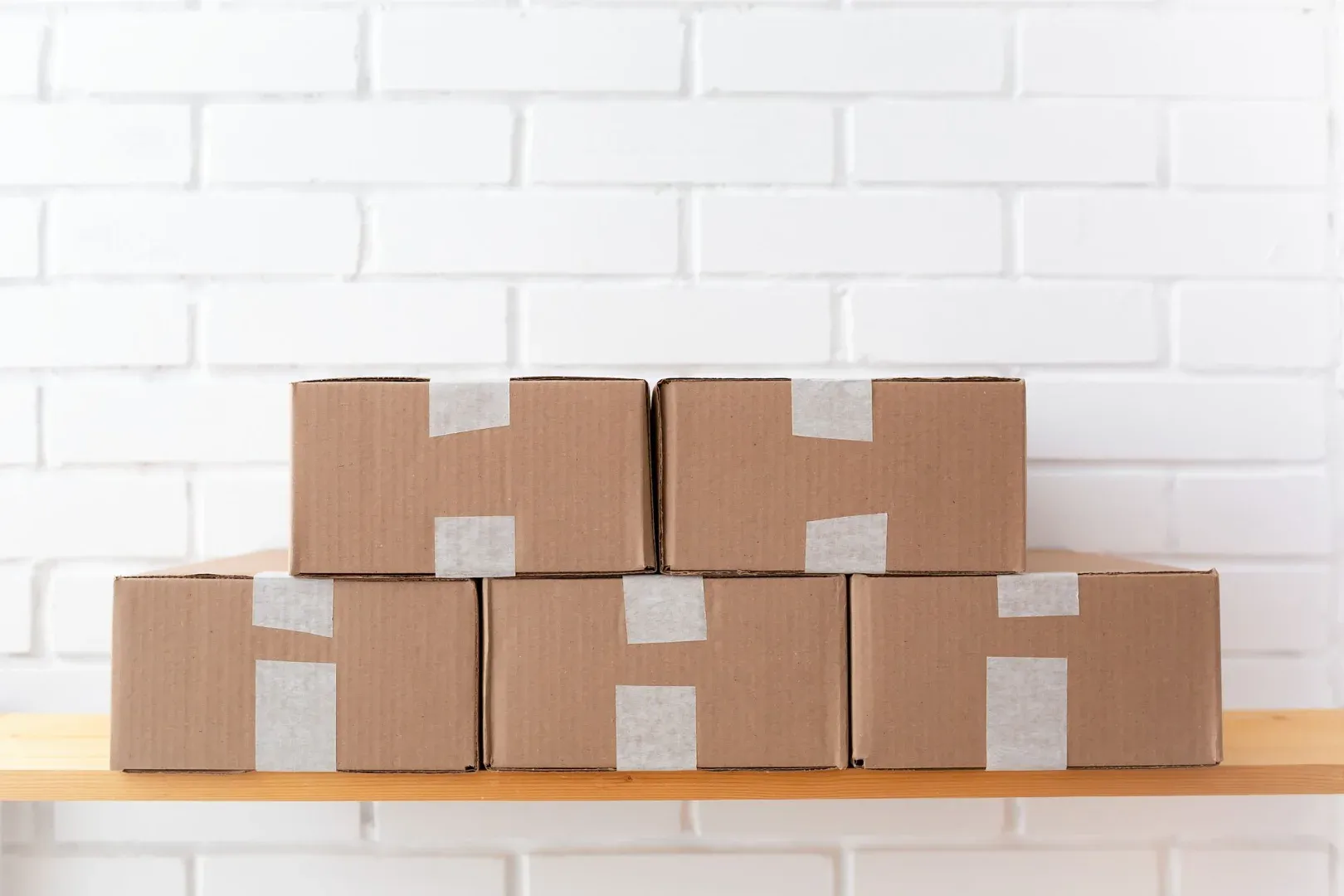 A neat stack of cardboard boxes taped and placed on a wooden shelf against a white brick wall.