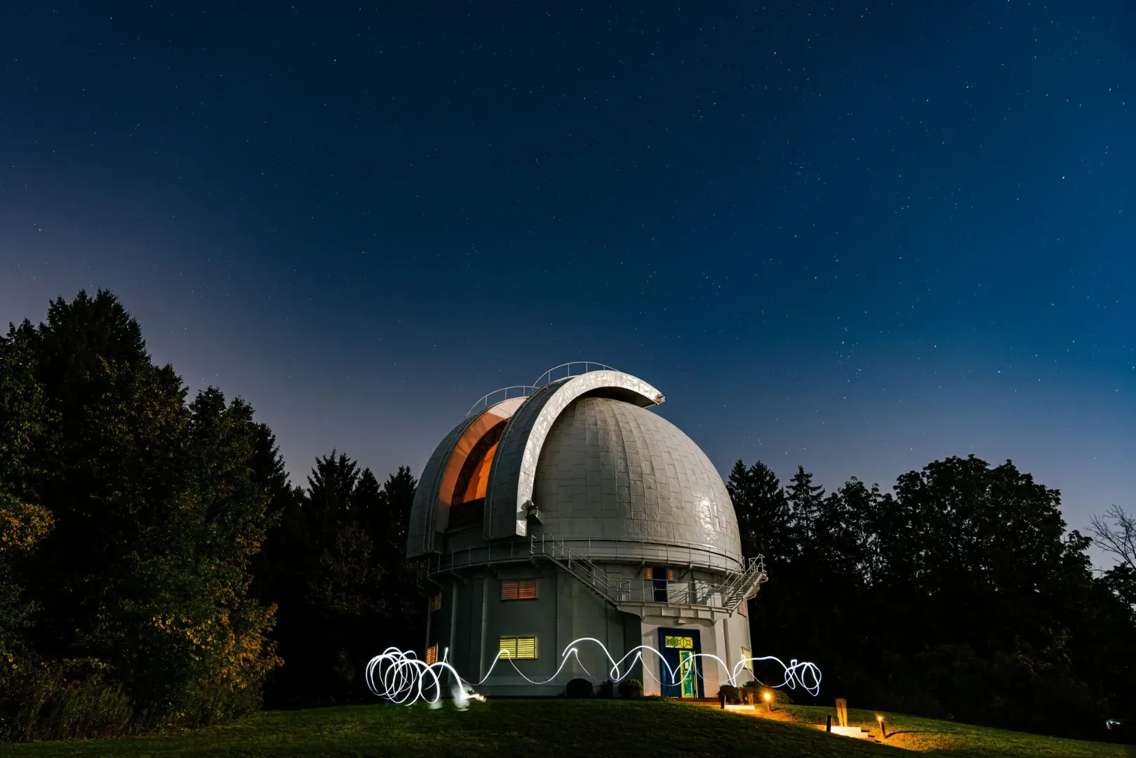 A scenic view of an observatory with a star-filled sky at night, surrounded by trees.