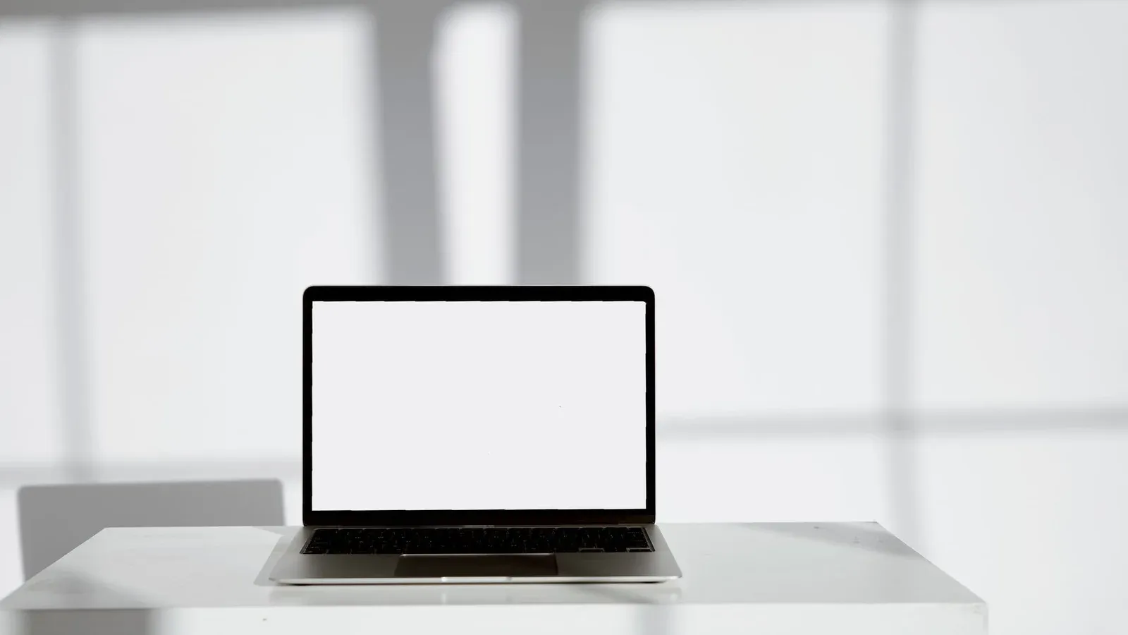 Blank laptop screen on a modern white desk in a bright, sunlit room.