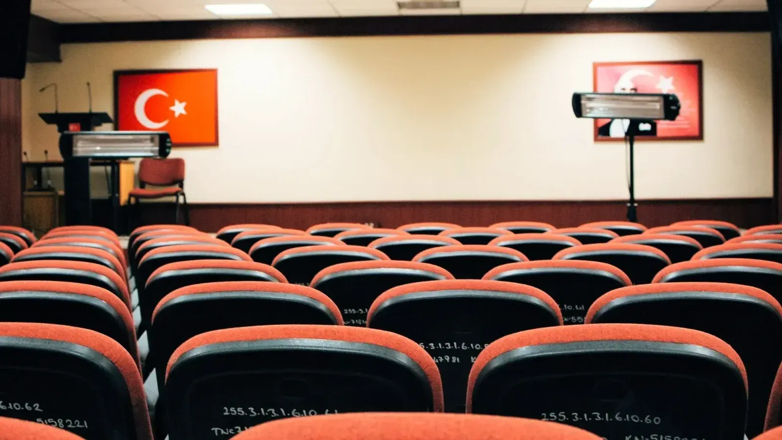 Una sala de conferencias vacía con asientos rojos, un escenario y banderas turcas, lista para una reunión o presentación.